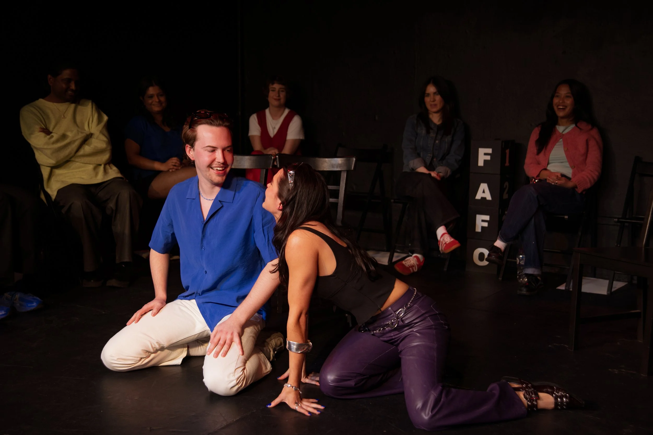 Two people are in a playful romantic pose on the floor, with the man kneeling and the woman leaning close to him, and several people are sitting on chairs in the background, watching and smiling.