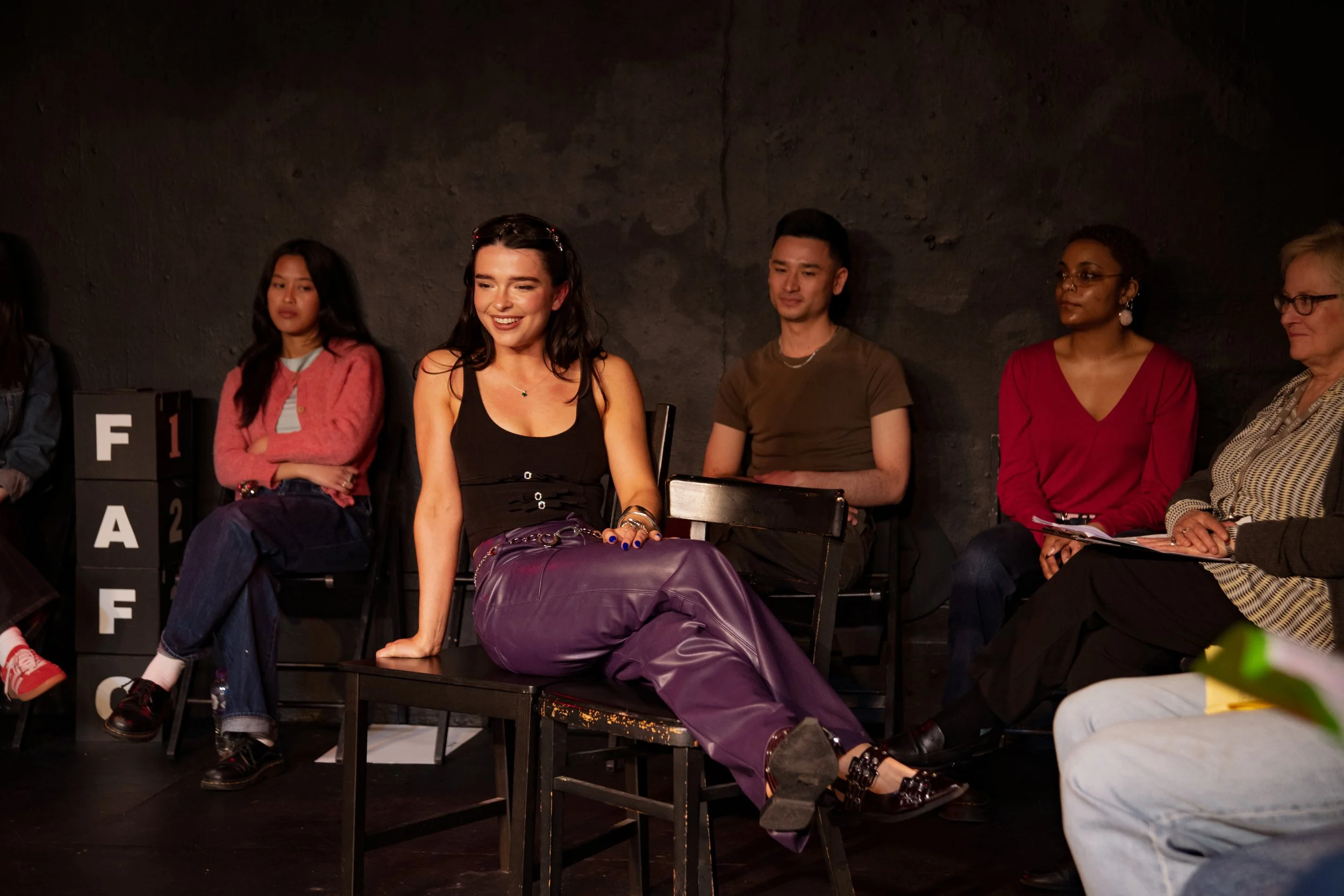 Group of diverse people sitting on chairs against dark wall, some looking towards the camera and others looking away, one person with legs crossed and one with feet resting on a table.
