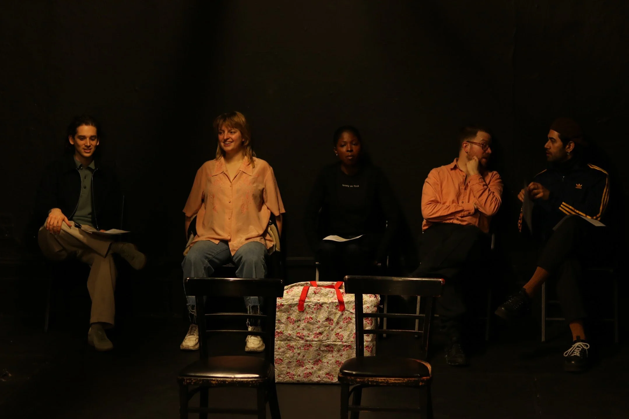 Five people are sitting in chairs against a black background, with a wrapped gift in the foreground.