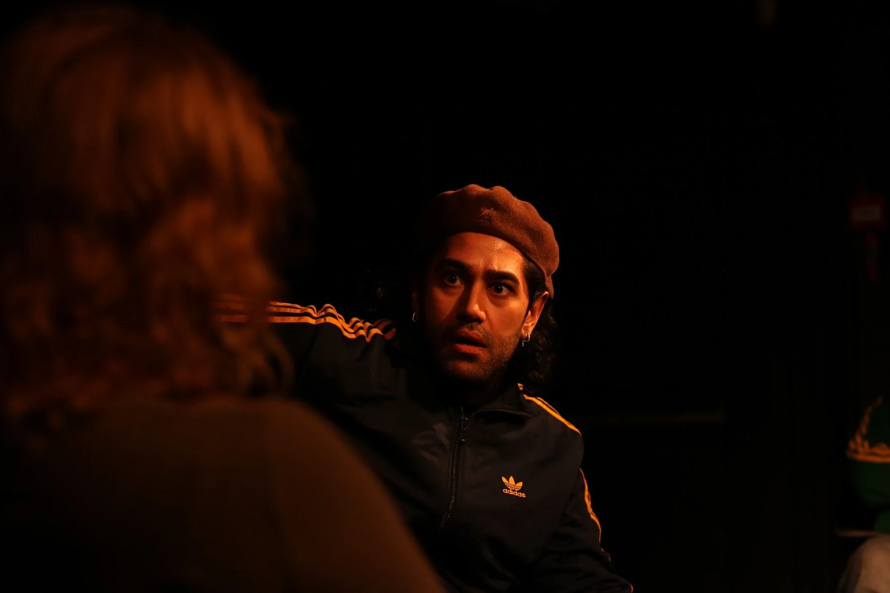A man with long dark hair, wearing earrings and a brown beret, looks at a woman with curly hair during a dark conversation at night.