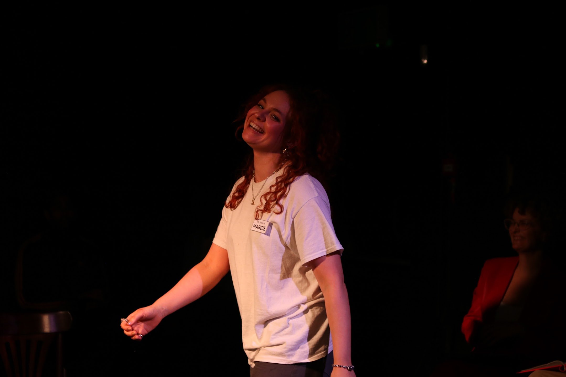 A young woman with curly red hair smiling, wearing a white T-shirt with a name tag, standing in a dark room.