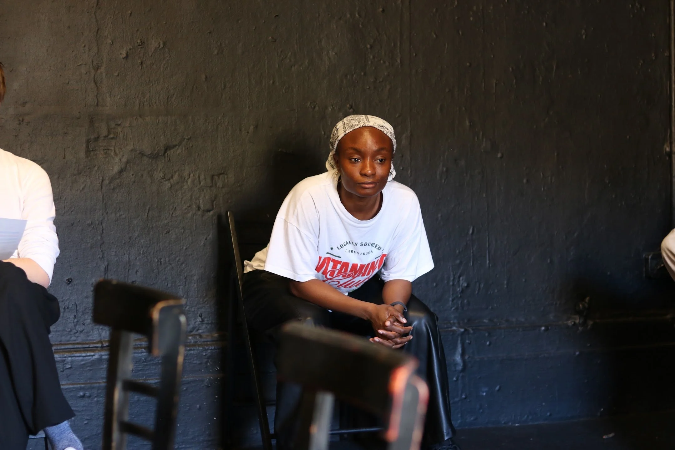 A young woman with dark skin and wearing a white bandana and a white T-shirt sitting against a black textured wall.