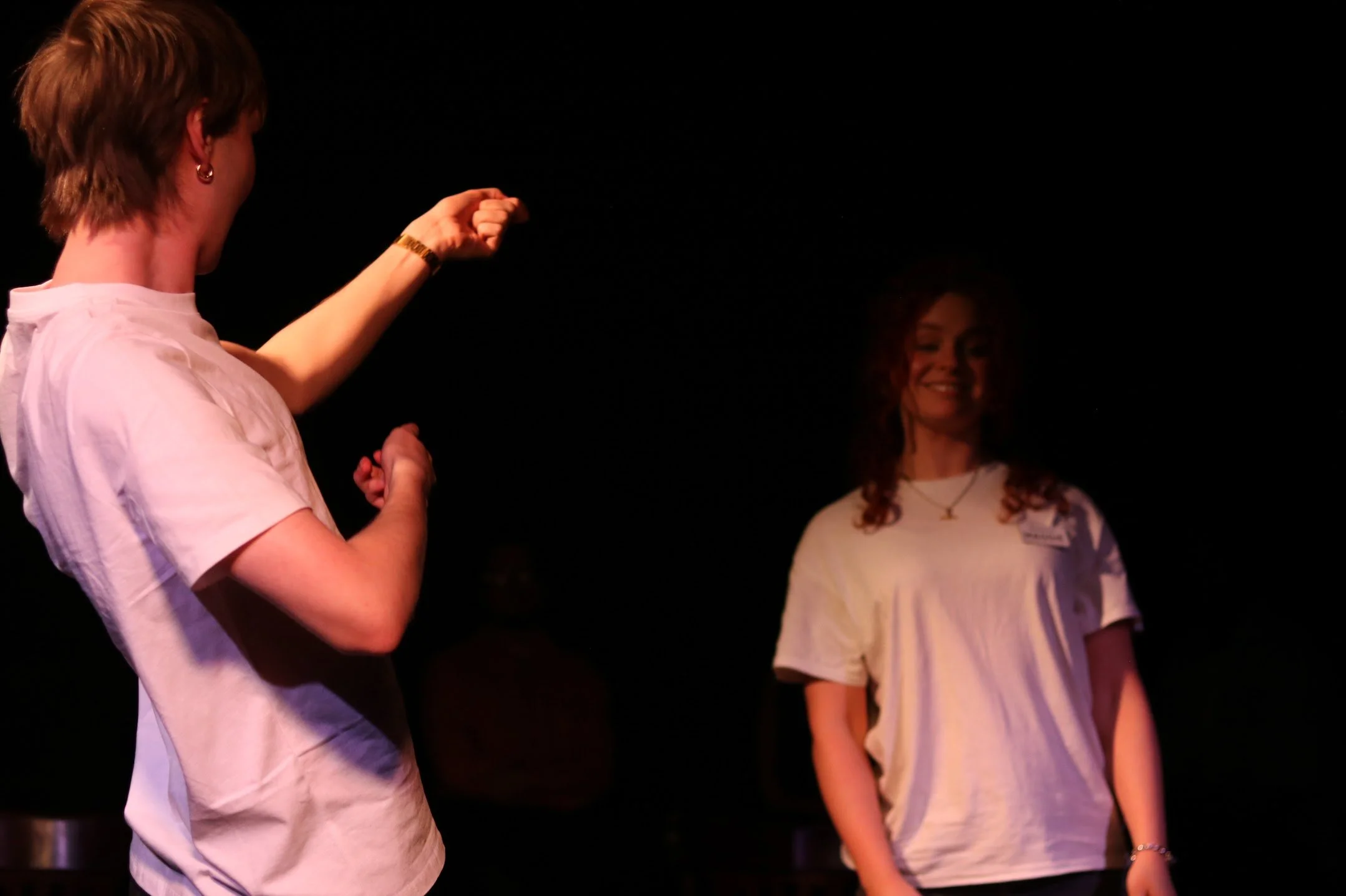Two women are standing on a dark stage. The woman on the left, with short hair and earrings, is gesturing with her fist, while the woman on the right, with long curly hair, is smiling and wearing a white top.
