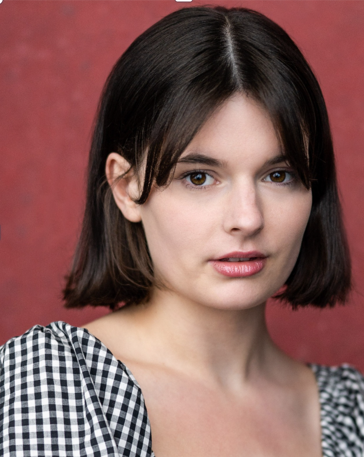 Close-up portrait of a young woman with shoulder-length dark brown hair, wearing a black and white checkered top, standing against a reddish background.