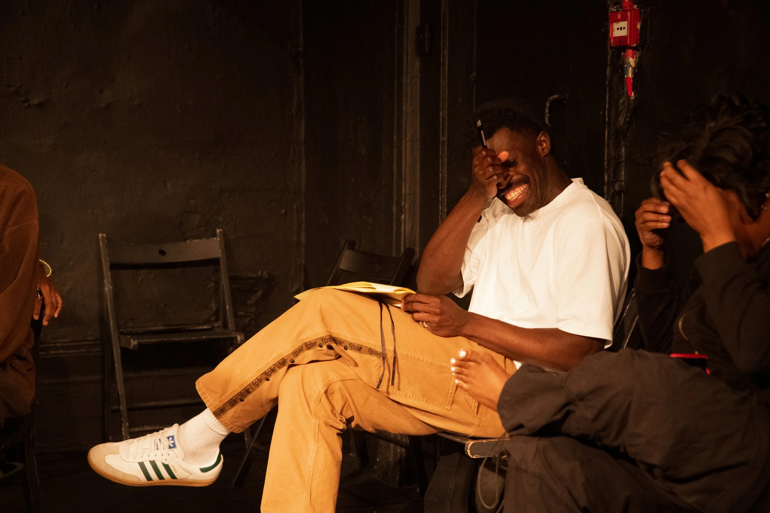 A man seated on a chair laughing with his hand on his forehead, wearing beige pants, a white shirt, and white sneakers with green stripes, with an orange bag on his lap, in a dark room with black walls and other chairs.