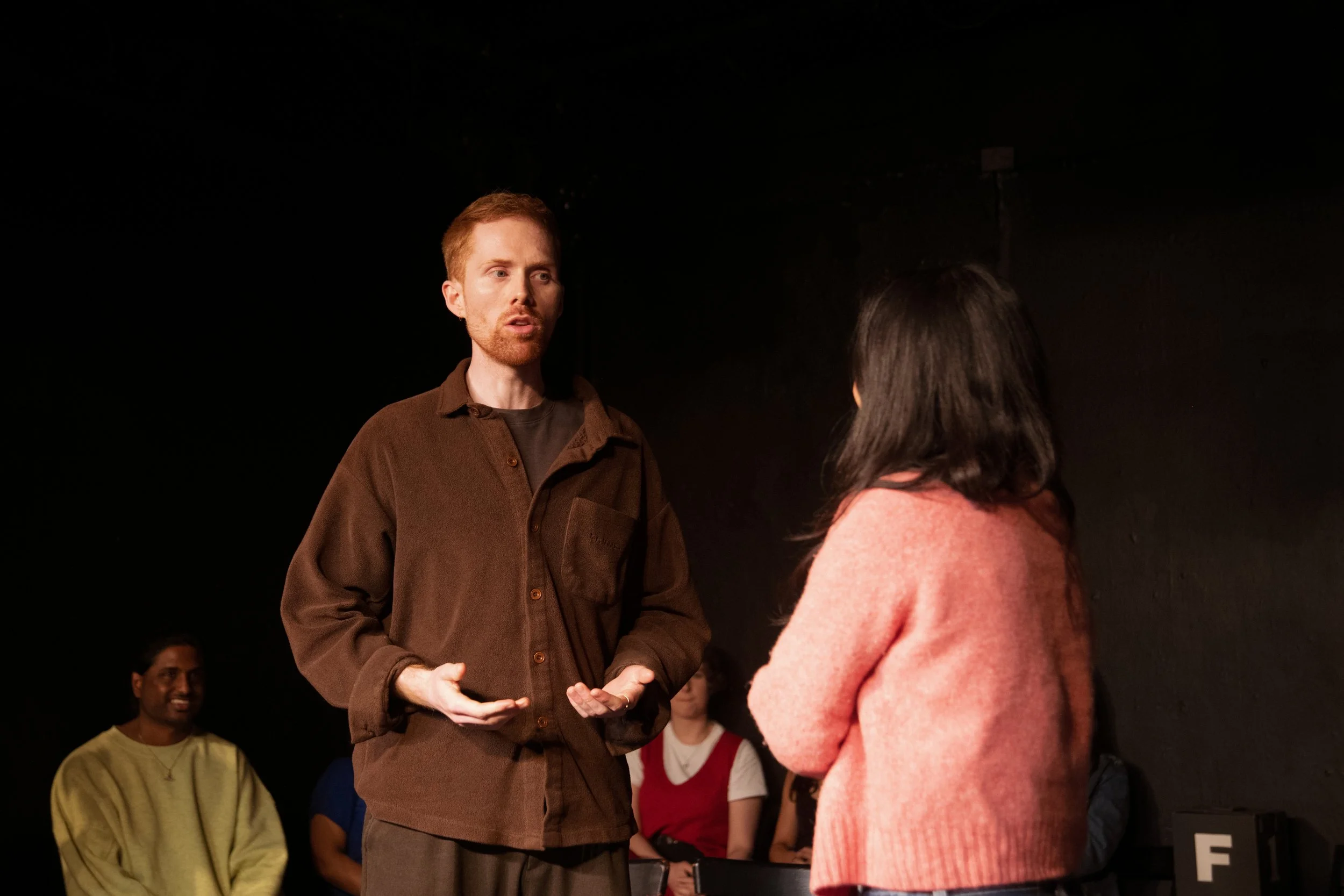 A man with red hair and beard wearing a brown jacket is speaking to a woman with dark hair wearing a pink sweater in a dark room. Several other people are visible in the background.
