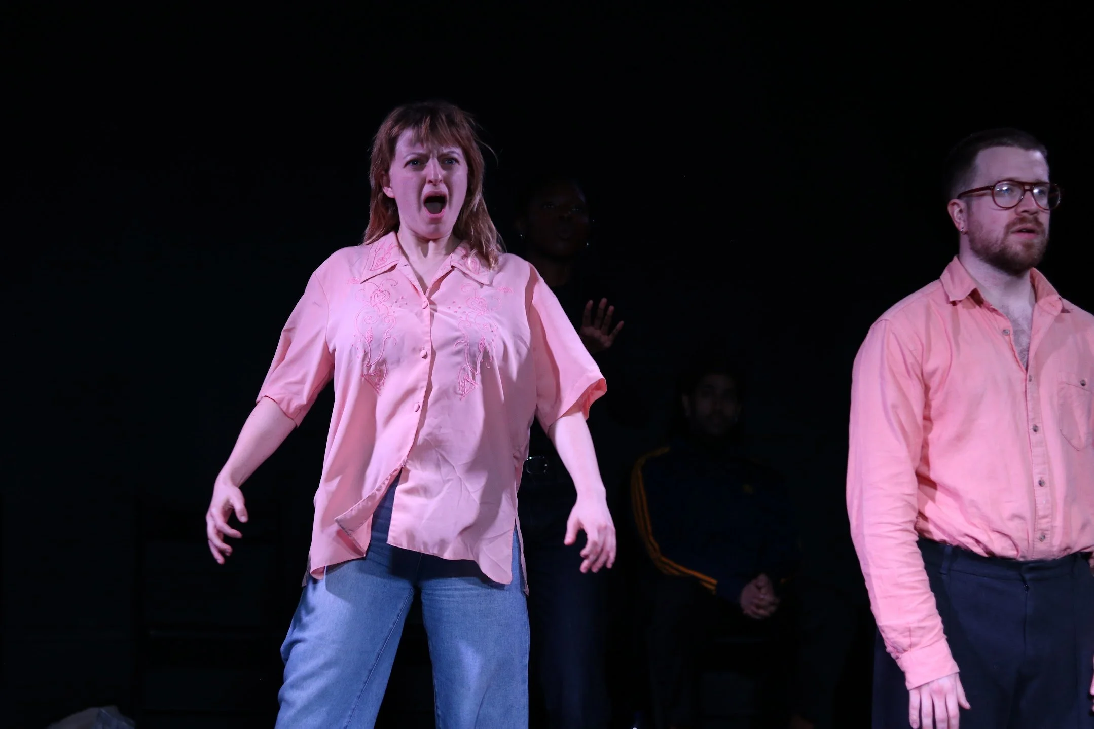 People standing on stage, wearing pink shirts, with one woman appearing to sing or shout, and a man next to her looking serious, in a dark background.