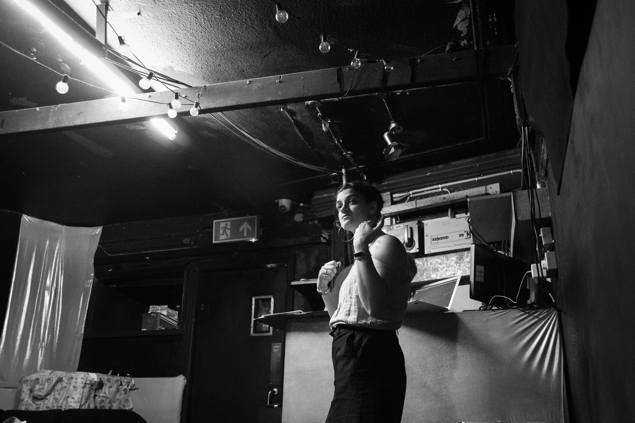 A woman standing in a room with a cluttered background, looking to the side, wearing a sleeveless top and earrings, with string lights hanging on the ceiling.