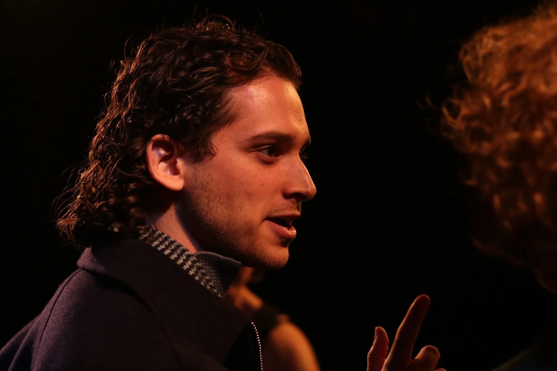 Profile of a man with wavy dark hair, wearing a dark coat and a checkered scarf, engaging in conversation with a woman with curly red hair, against a dark background.