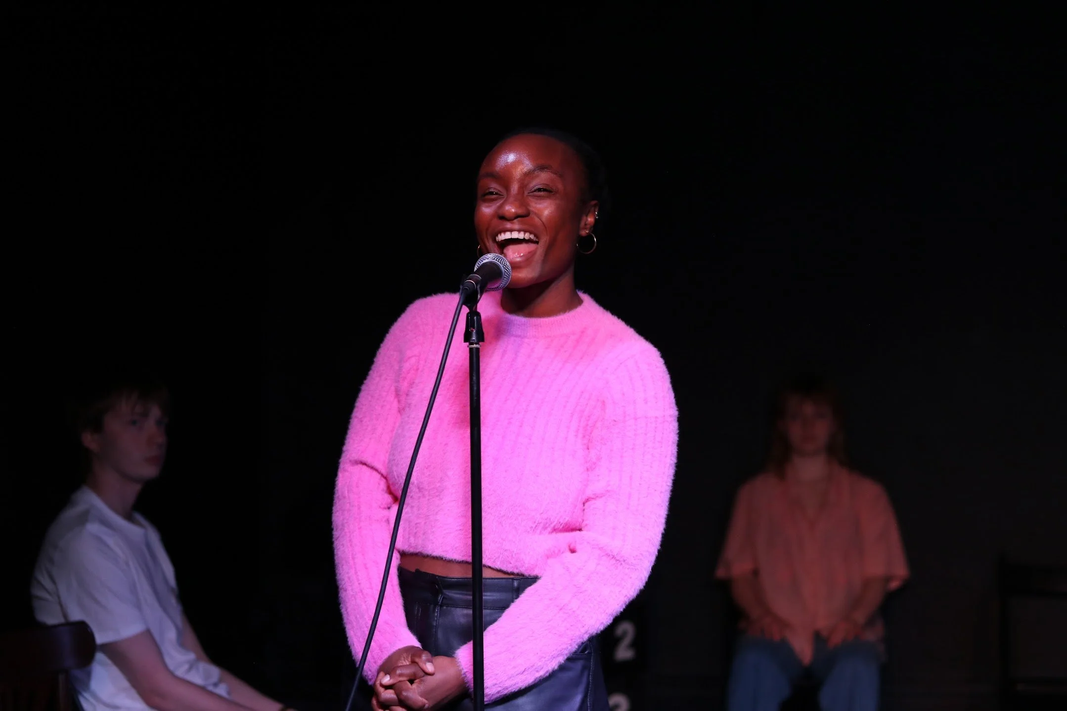 A woman wearing a pink fuzzy sweater and black pants speaking into a microphone on stage with a black background. There are three out-of-focus people in the background.
