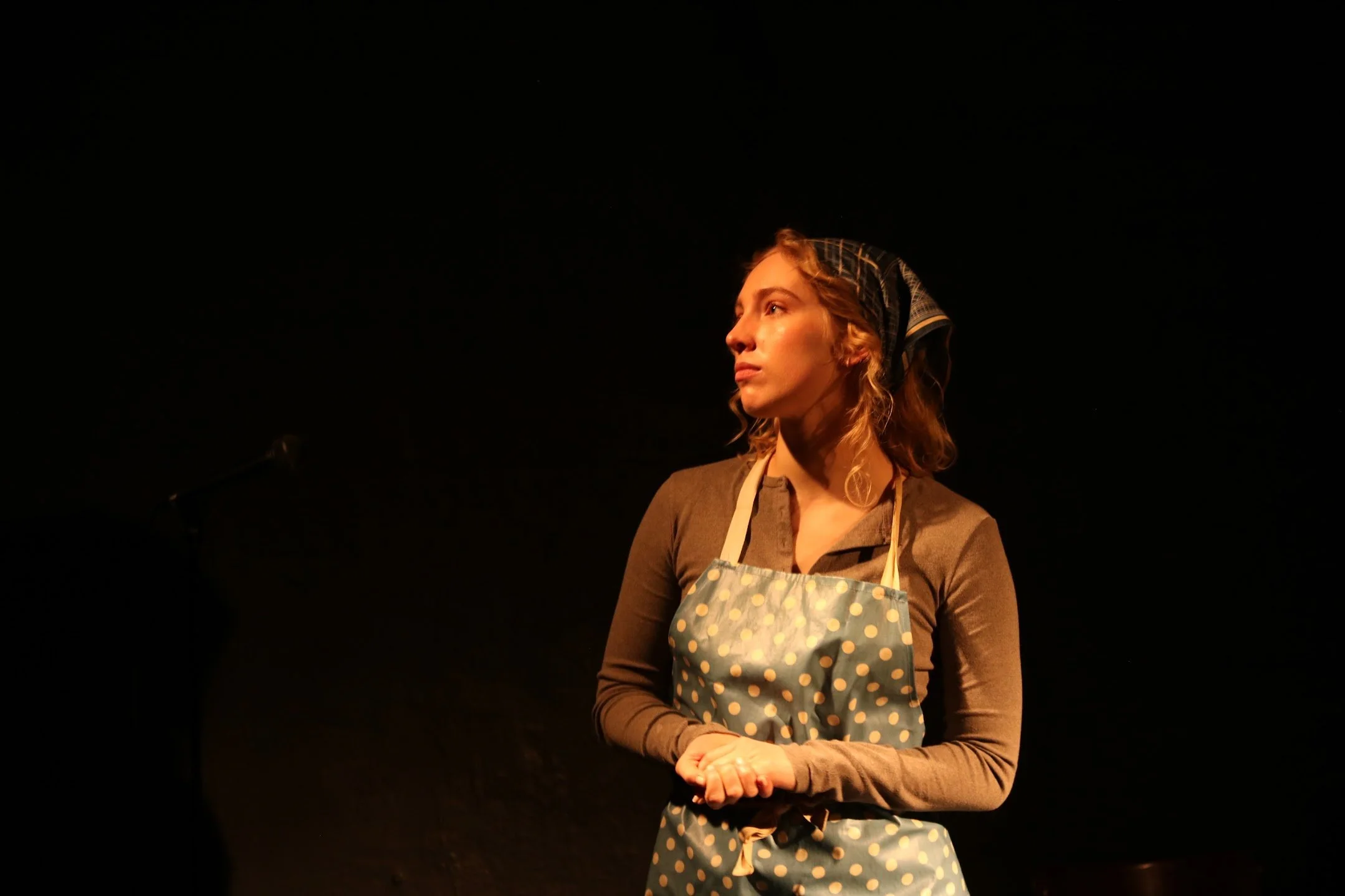 A woman with curly hair wearing a striped headscarf, a brown long-sleeve shirt, and a blue apron with yellow polka dots, standing against a dark background.