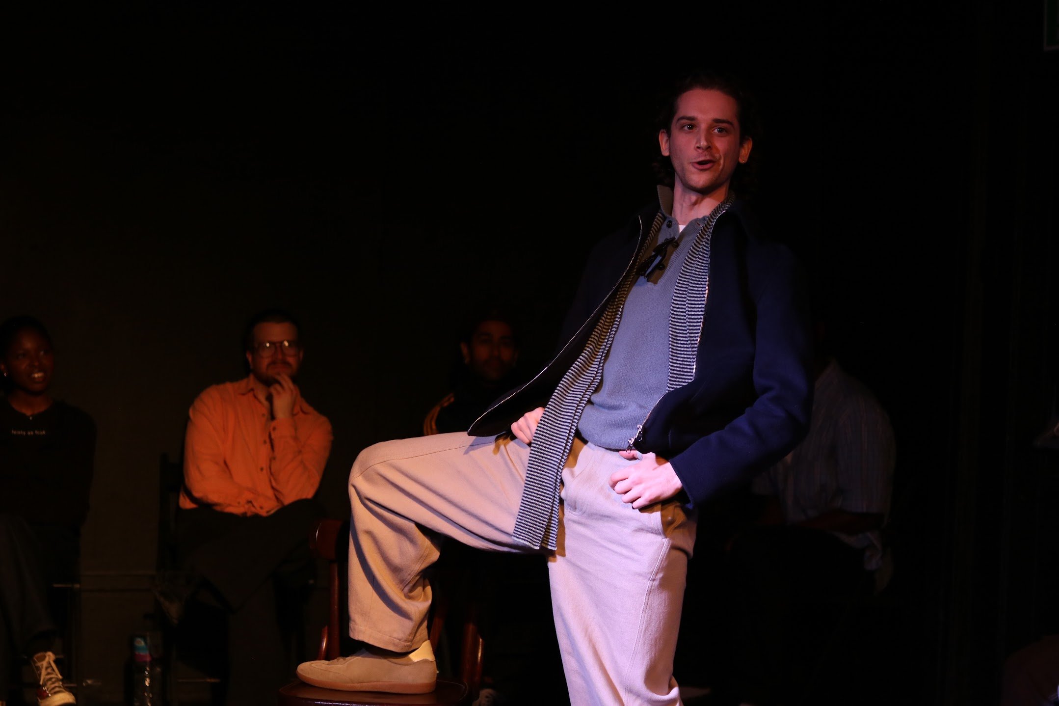 A young man with dark hair sitting on a stool on stage, speaking or singing, dressed in a gray shirt, black jacket, and beige pants. Four people are in the background, listening, with dark backgrounds and stage lighting.