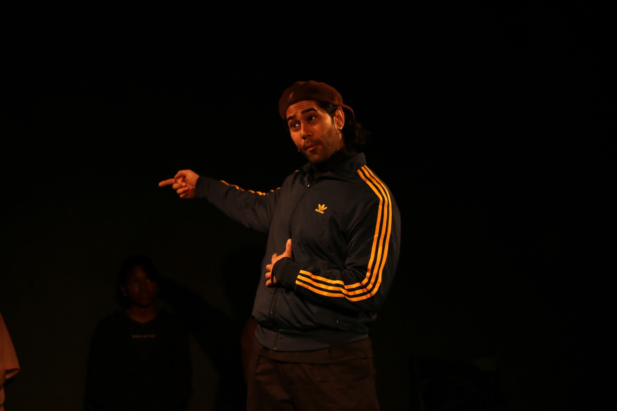 A man with dark hair and a beard, wearing a black Adidas jacket with yellow stripes and a backward baseball cap, is pointing with his right hand while looking to his left in a dark environment.