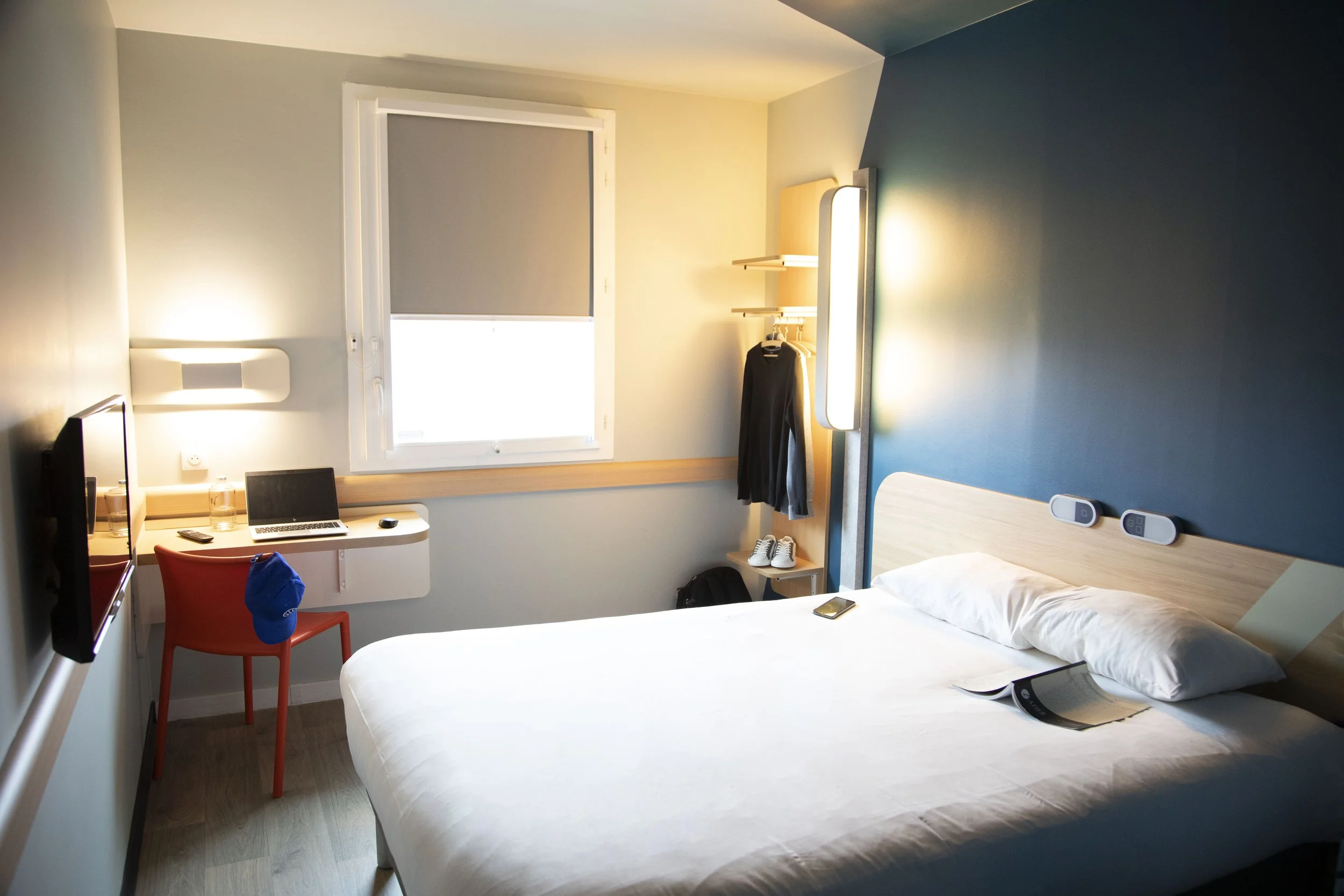 Chambre d'hôtel avec un lit, un bureau avec un ordinateur portable, une lampe, une chaise orange, une fenêtre avec un store, et des éléments de décoration modernes.