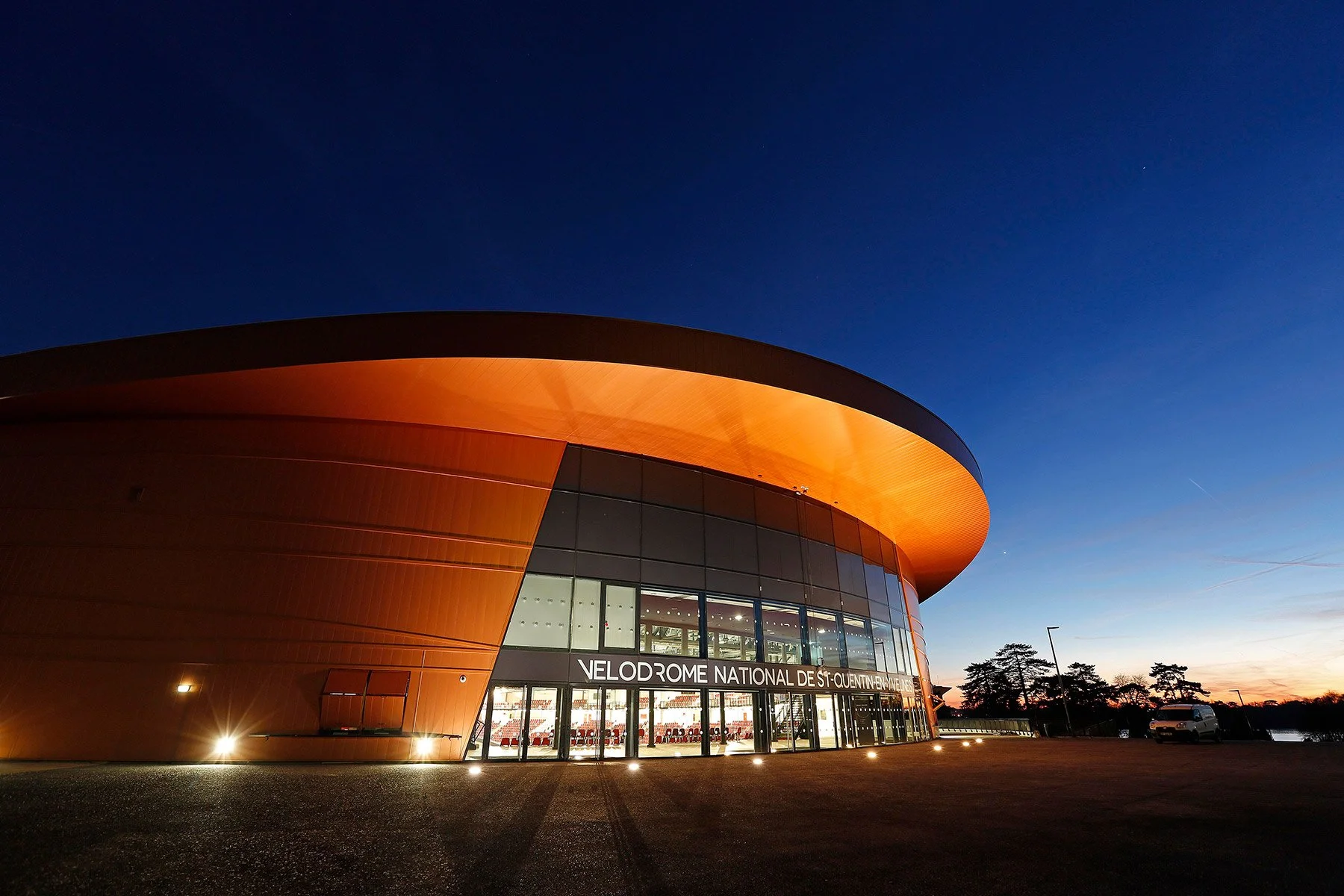 Nighttime view of the Velodrome National de Saint-Quentin-en-Yvelines, a modern, curved building with orange exterior and large glass windows, illuminated from inside.