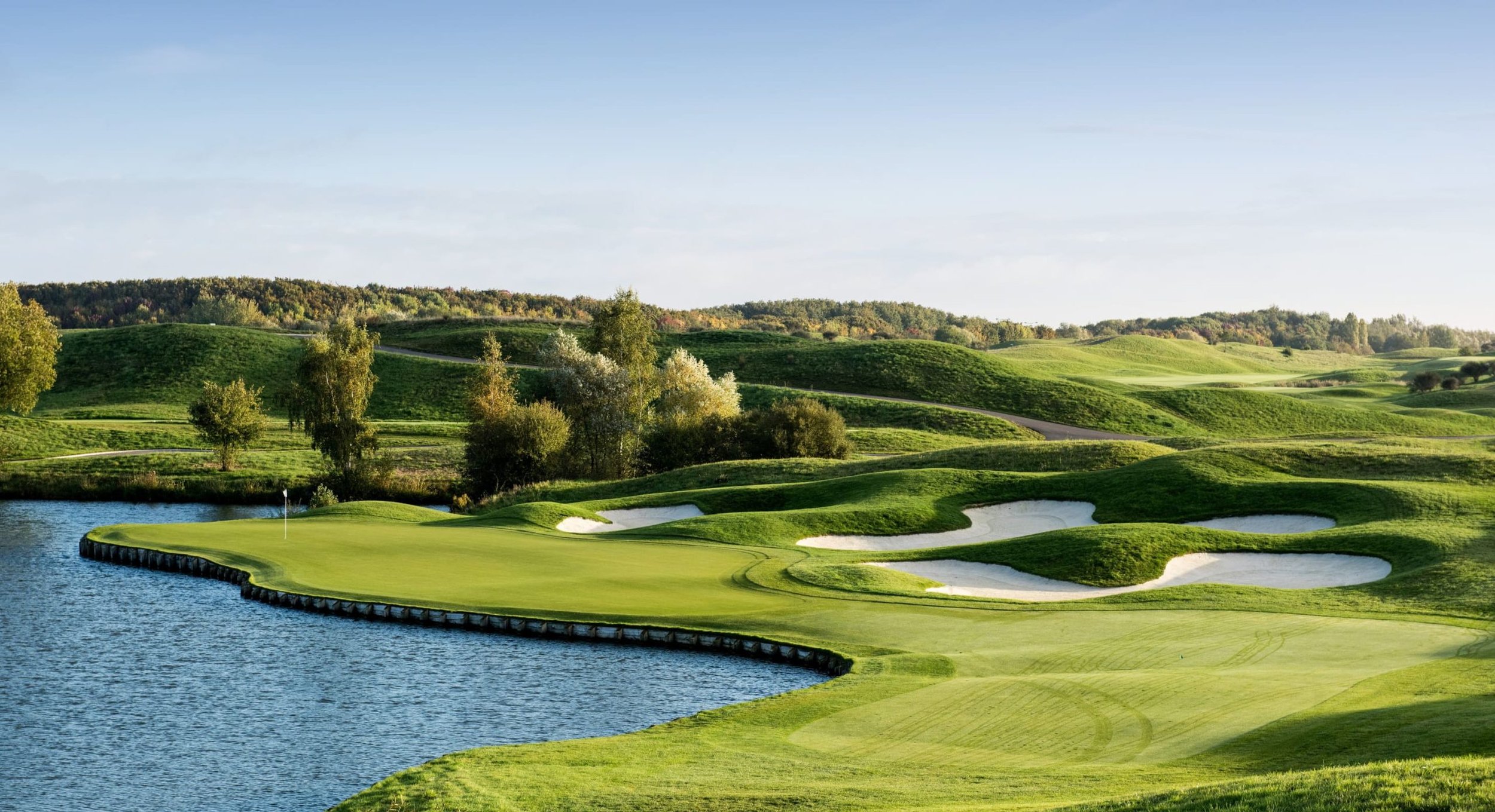 A golf course with a green, sand bunkers, water hazard, and rolling hills in the background on a sunny day.