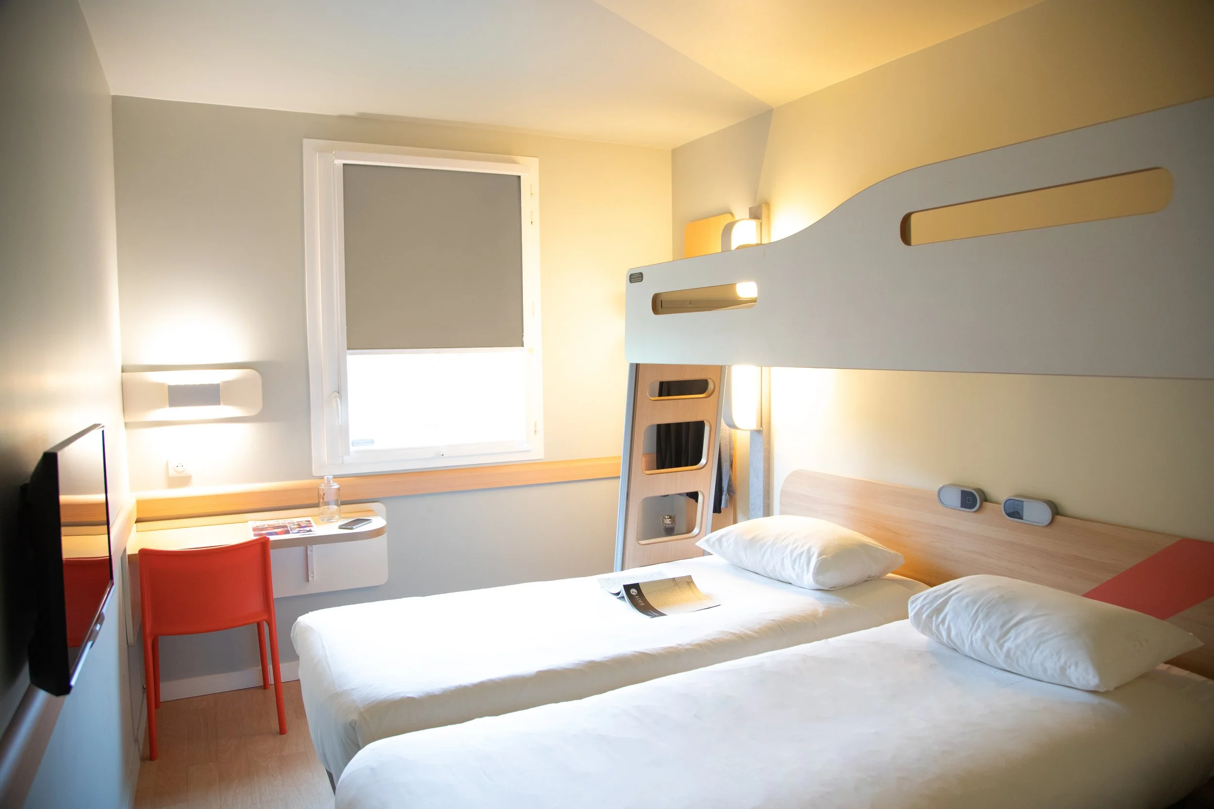 Chambre d'hôtel moderne avec deux lits simples, une fenêtre avec volet, une lampe murale, un petit bureau avec une chaise rouge, et des équipements électriques à côté du lit.
