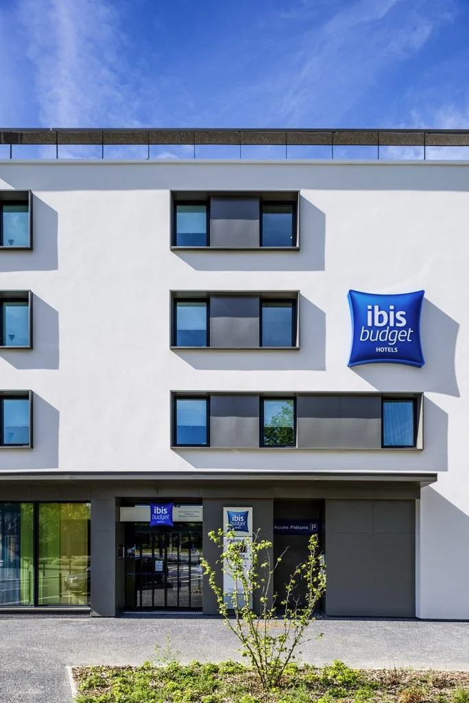 Façade d'un hôtel Ibis Budget avec un signe en tissu bleu affichant le logo de l'hôtel. La façade est blanche avec des fenêtres rectangulaires et le bâtiment est sous un ciel bleu.