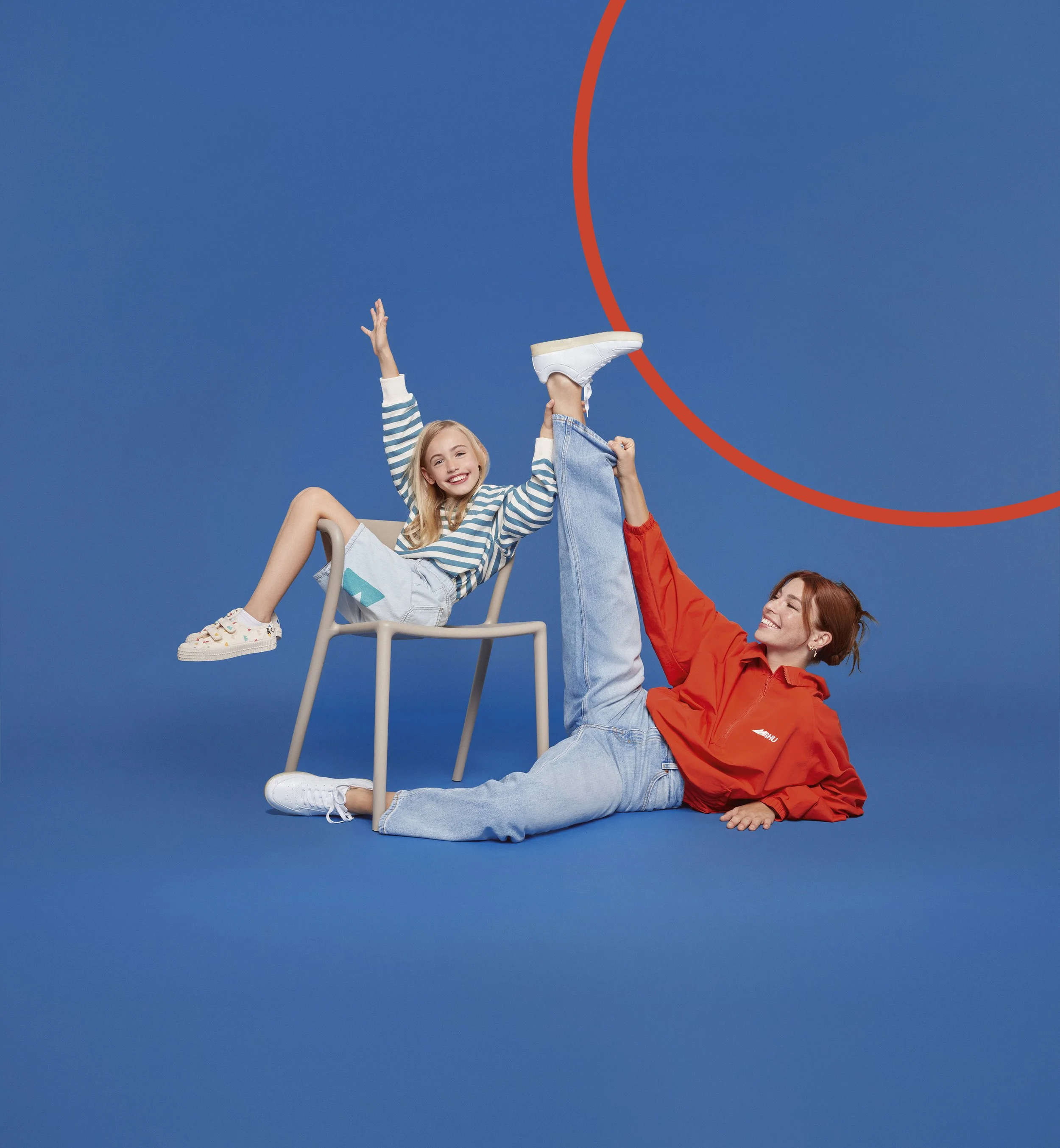 Une femme souriante, en veste orange, aide une jeune fille à faire un signe de la main tout en étant assise sur une chaise. La jeune fille est vêtue d'un pull à rayures bleues et blanches, d'un short en jean, et porte des baskets colorées. Elles sont contre un fond bleu avec une ligne courbe orange.