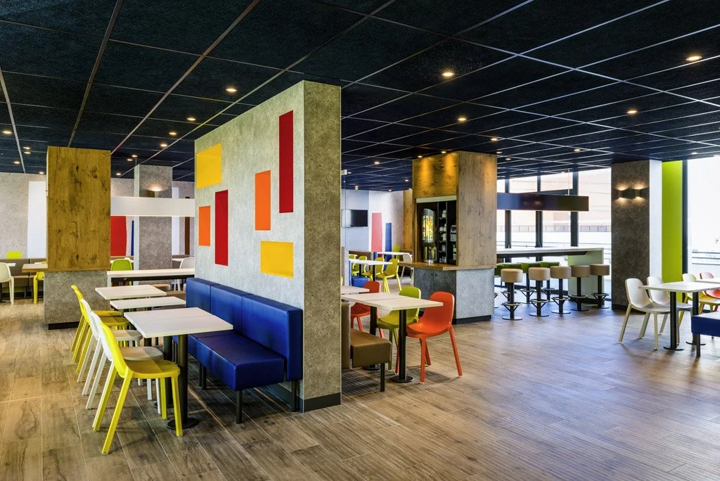 Salle de restaurant moderne avec tables et chaises colorées, murs décorés avec des formes géométriques, fenêtres larges laissant entrer la lumière naturelle.