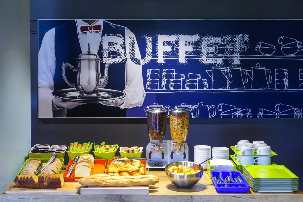 Buffet de petit-déjeuner avec plusieurs pains, céréales, fruits, yaourts, tasses, cuillères, et contenants pour boissons chaudes