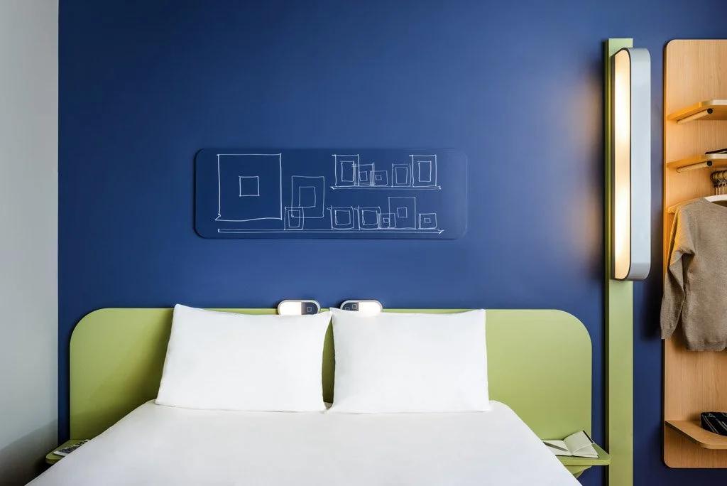 Camera con letto matrimoniale, cuscini bianchi, parete blu con una mensola a muro bianca e illuminazione moderna sul lato destro.