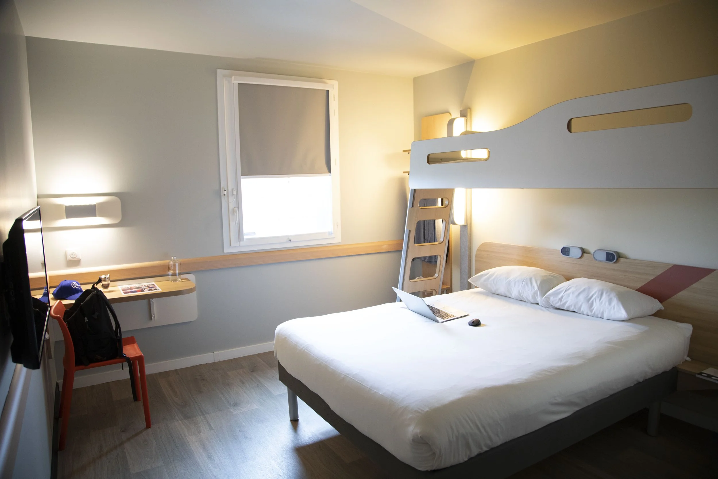 Chambre d'hôtel moderne avec lit double, bureau, et lit superposé au fond, fenêtre avec volet, et éléments d'éclairage