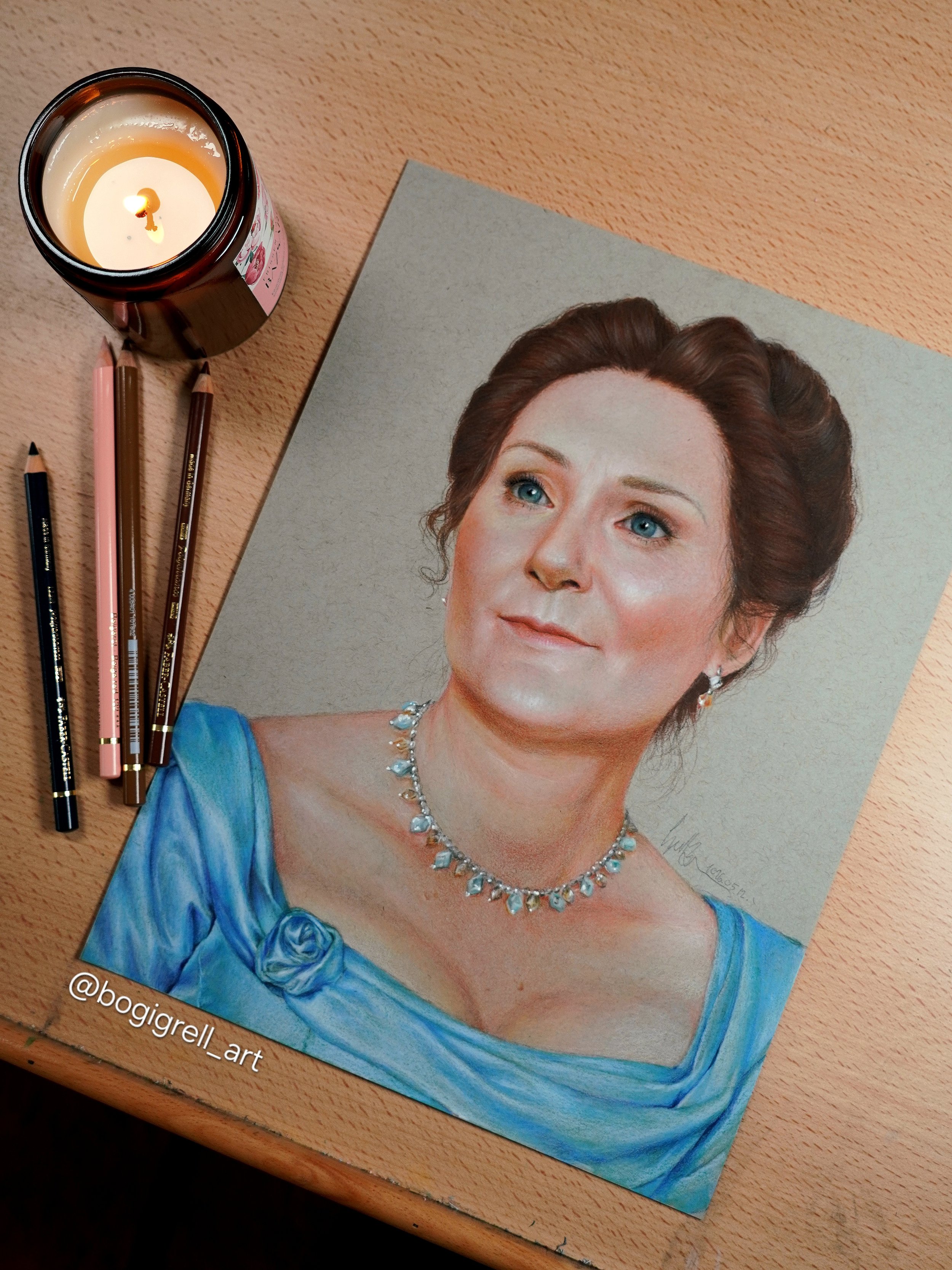 A realistic portrait drawing of a woman with blue eyes, brown hair styled up, wearing a blue dress with a floral detail, and a diamond necklace, placed on a wooden surface next to three colored pencils and a lit candle. The Woman is Violet Bridgerton