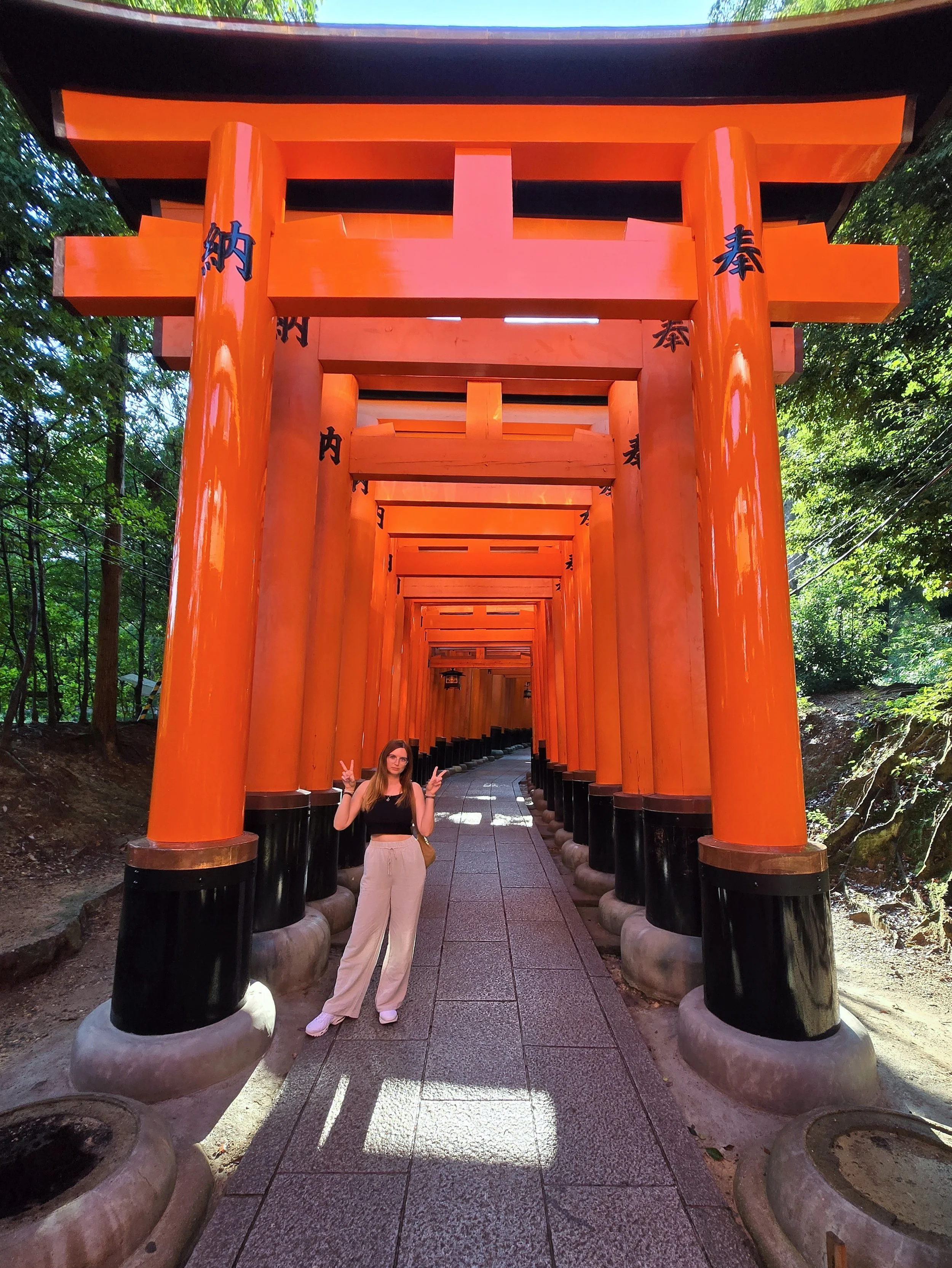 A woman standing in front of a series of bright orange torii gates at a Japanese shrine, making a peace sign with both hands.