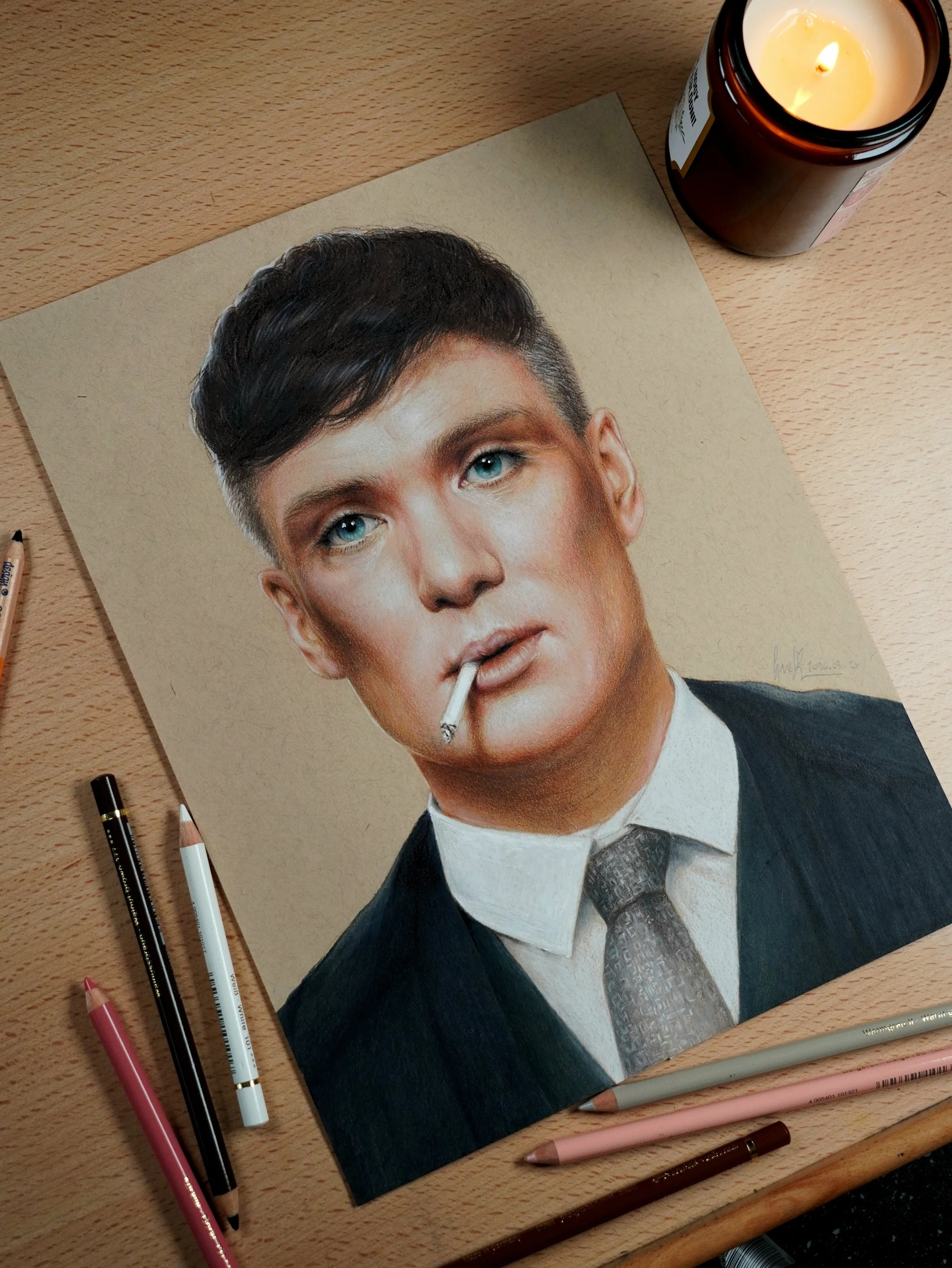 A realistic colored pencil drawing of a person with short dark hair, blue eyes, and fair skin, wearing a black suit, white shirt, and patterned gray tie, holding a lit cigarette in mouth. The drawing is on a beige textured paper on a wooden surface. Various pencils and markers are placed around it, and a lit candle in a brown jar is nearby.