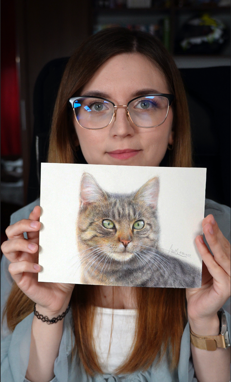 A young woman with glasses holding a detailed colored pencil drawing of a gray and brown tabby cat, in a room with shelves in the background.