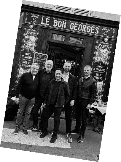 Five men standing outside a restaurant called Le Bon Georges, posing for a photo. The restaurant has a signboard with menu items written on it.