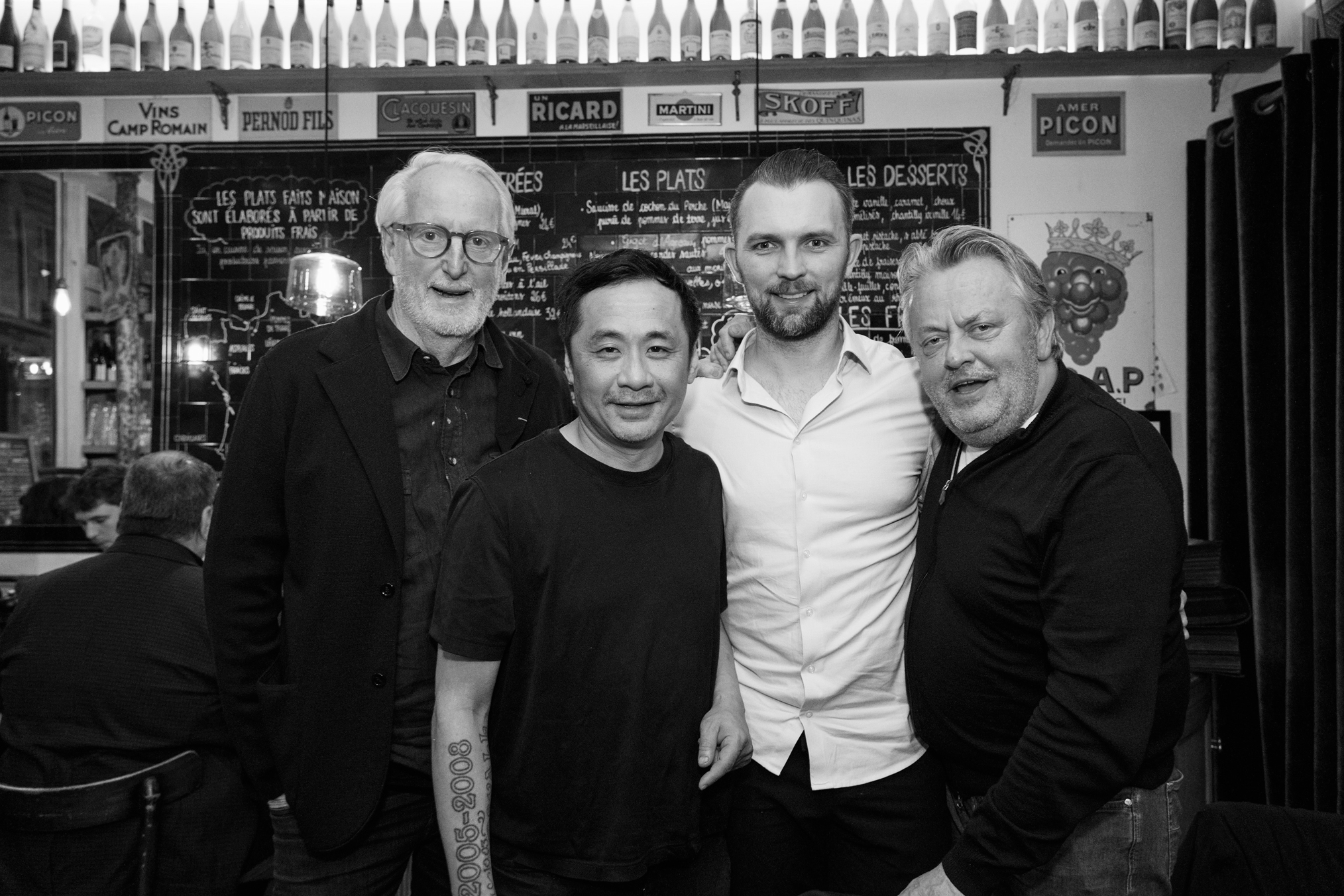 Four men standing together inside a restaurant, smiling at the camera, with a chalkboard menu and wine bottles in the background.