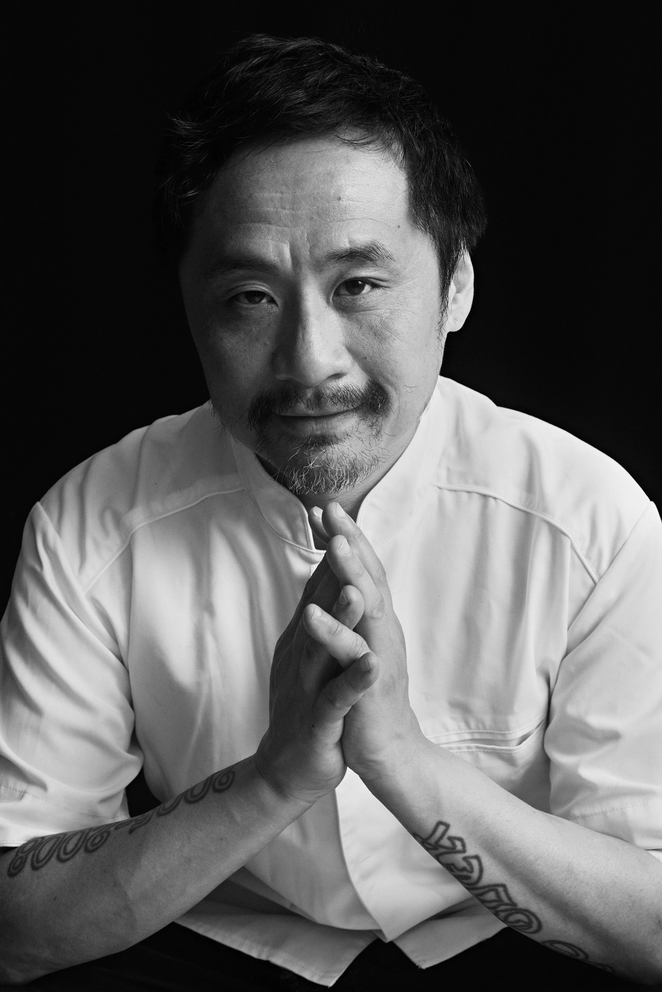 Black and white portrait of a man with short dark hair, mustache, and goatee, wearing a chef's jacket, with hands pressed together in a prayer-like gesture, against a dark background.