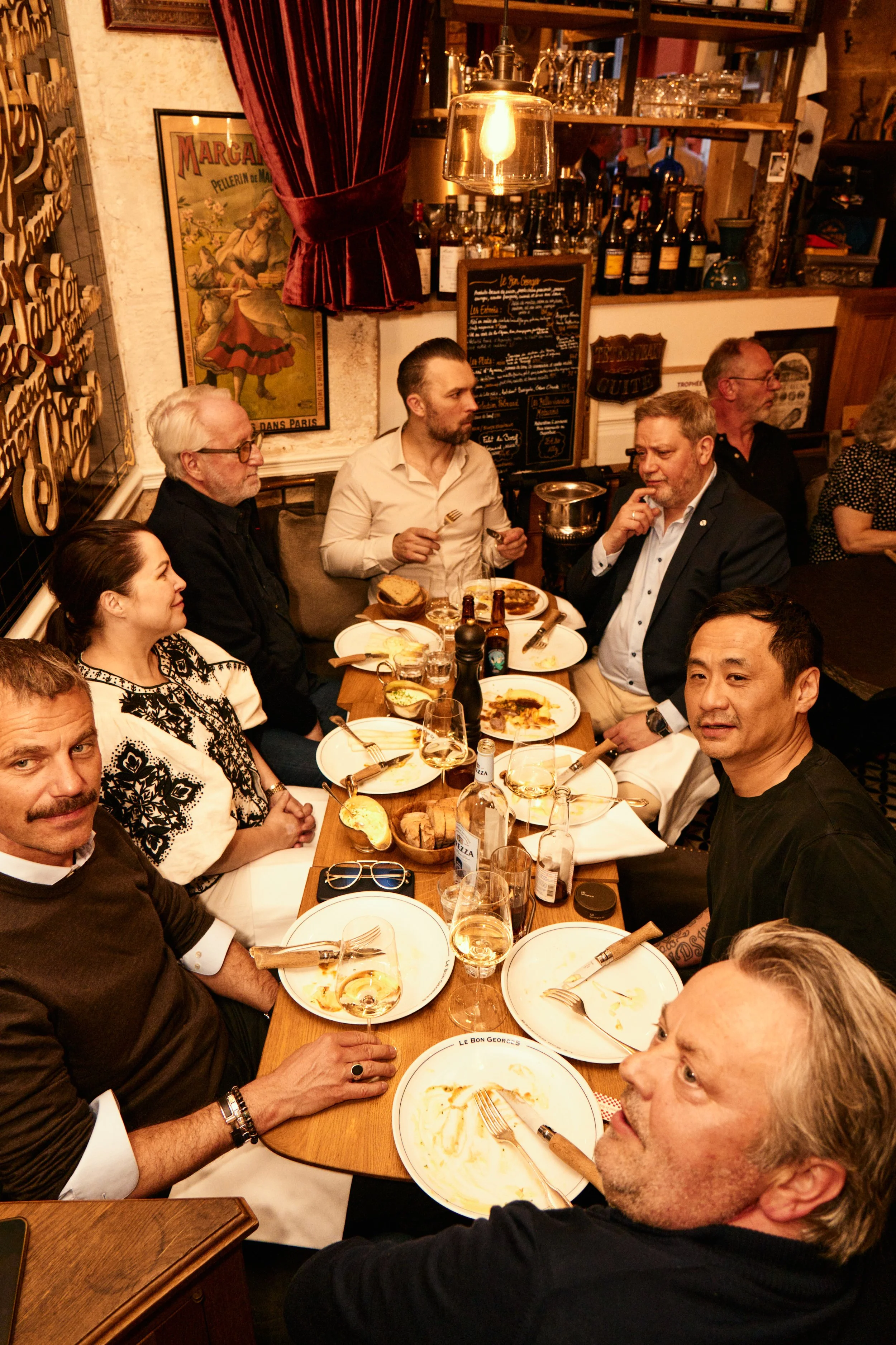 A group of people dining at a restaurant, with plates of food and drinks on the table. The background features a bar with bottles, glasses, and a chalkboard menu, and various decorative items on the walls.