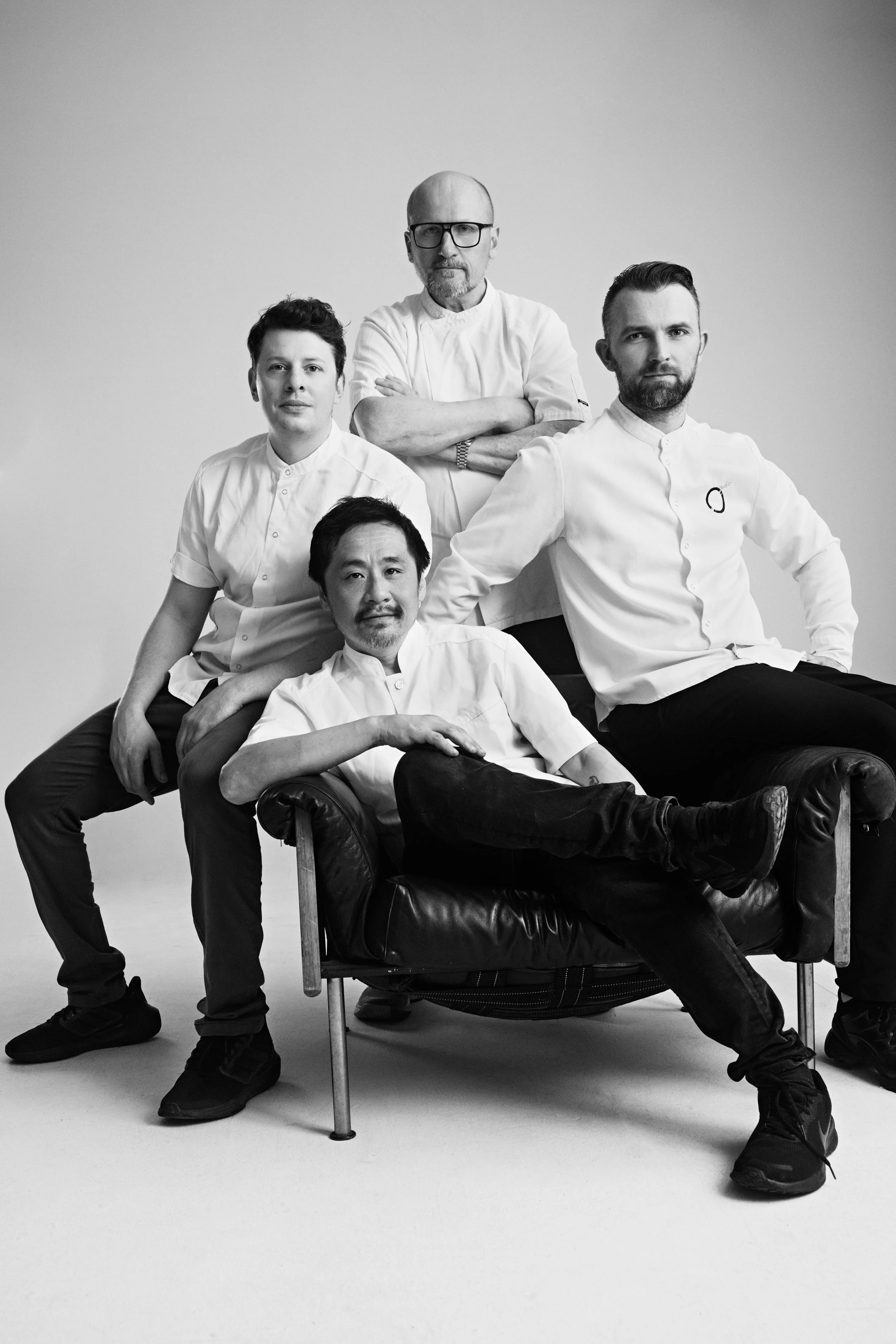 Black and white photo of five people, possibly chefs, posing together against a plain background. One person is sitting on a beanbag chair in the foreground, while the others stand or sit around him.