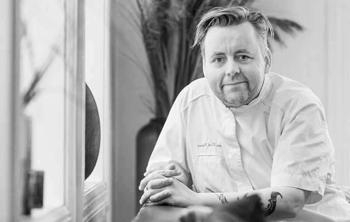 Man with a beard wearing a chef's coat, resting his hands on a counter, in a kitchen with plants in the background.