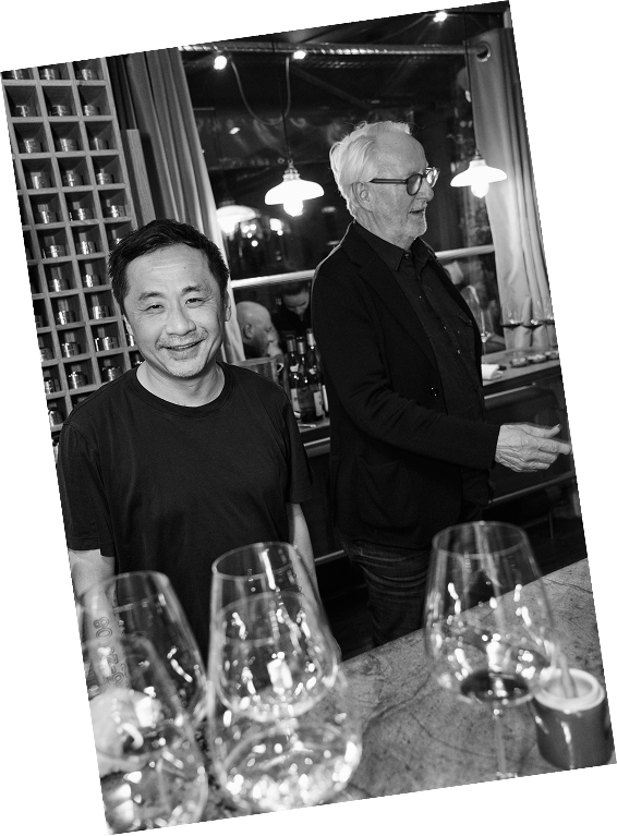 Two men standing in a modern bar with wine glasses in the foreground. One man is Asian, smiling, and the other is Caucasian with white hair, wearing glasses, looking to the side. The bar has bottles on shelves, wine glasses, and warm lighting.