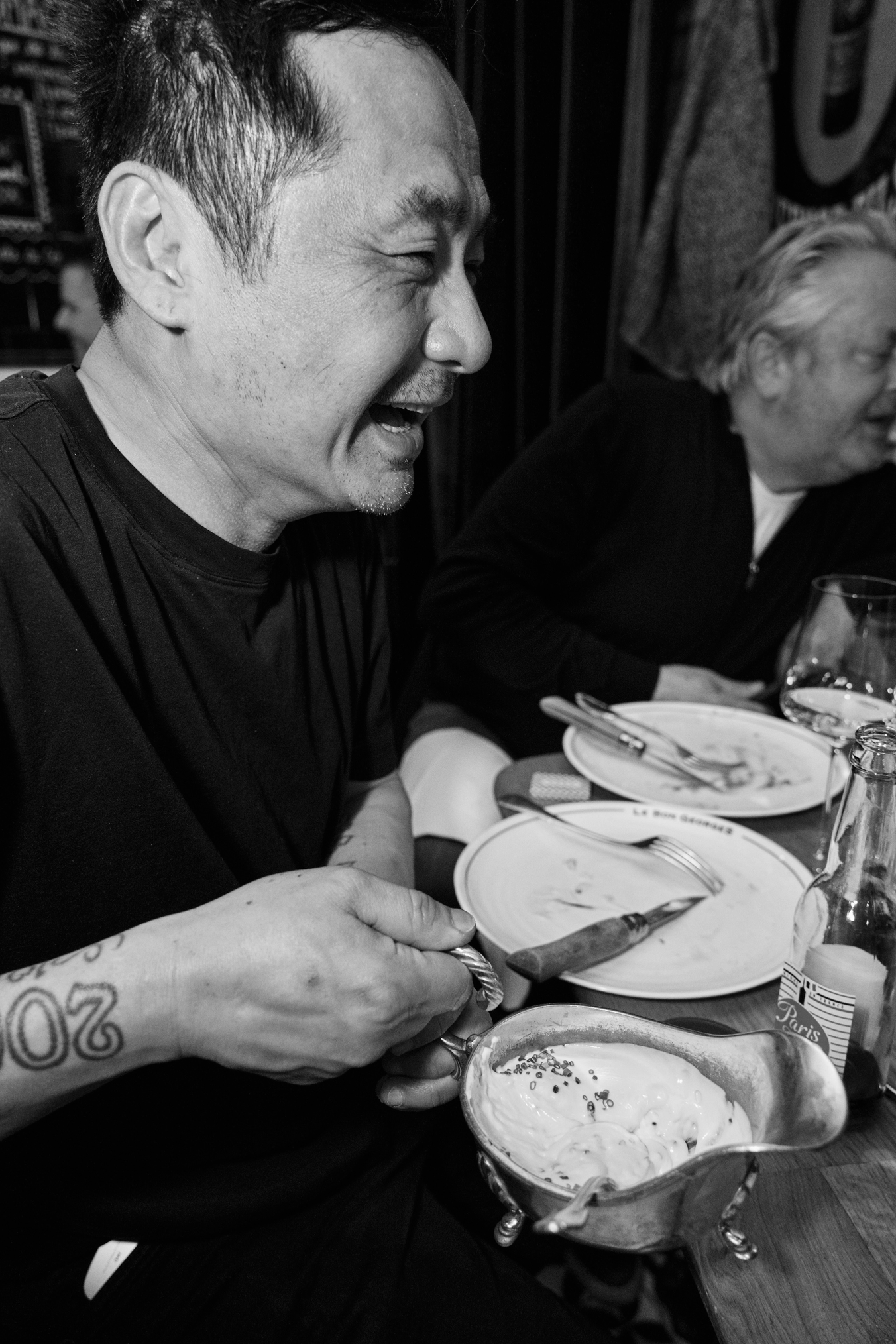 A man with short dark hair and facial stubble, laughing with eyes closed, sits at a dining table with a woman. They are enjoying a meal with various dishes, including a bowl of creamy soup topped with herbs. The man is holding a spoon and knife, and there are plates, glasses, and utensils on the table.