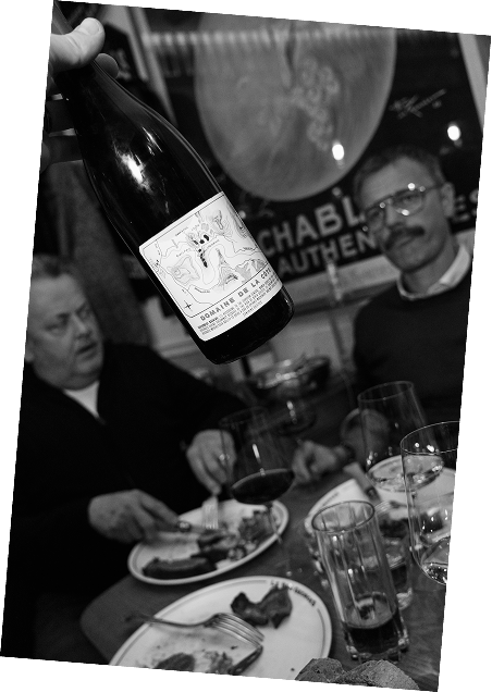 A person holding a wine bottle over a dinner table with two men sitting, glasses of wine, and plates of food in a restaurant setting.