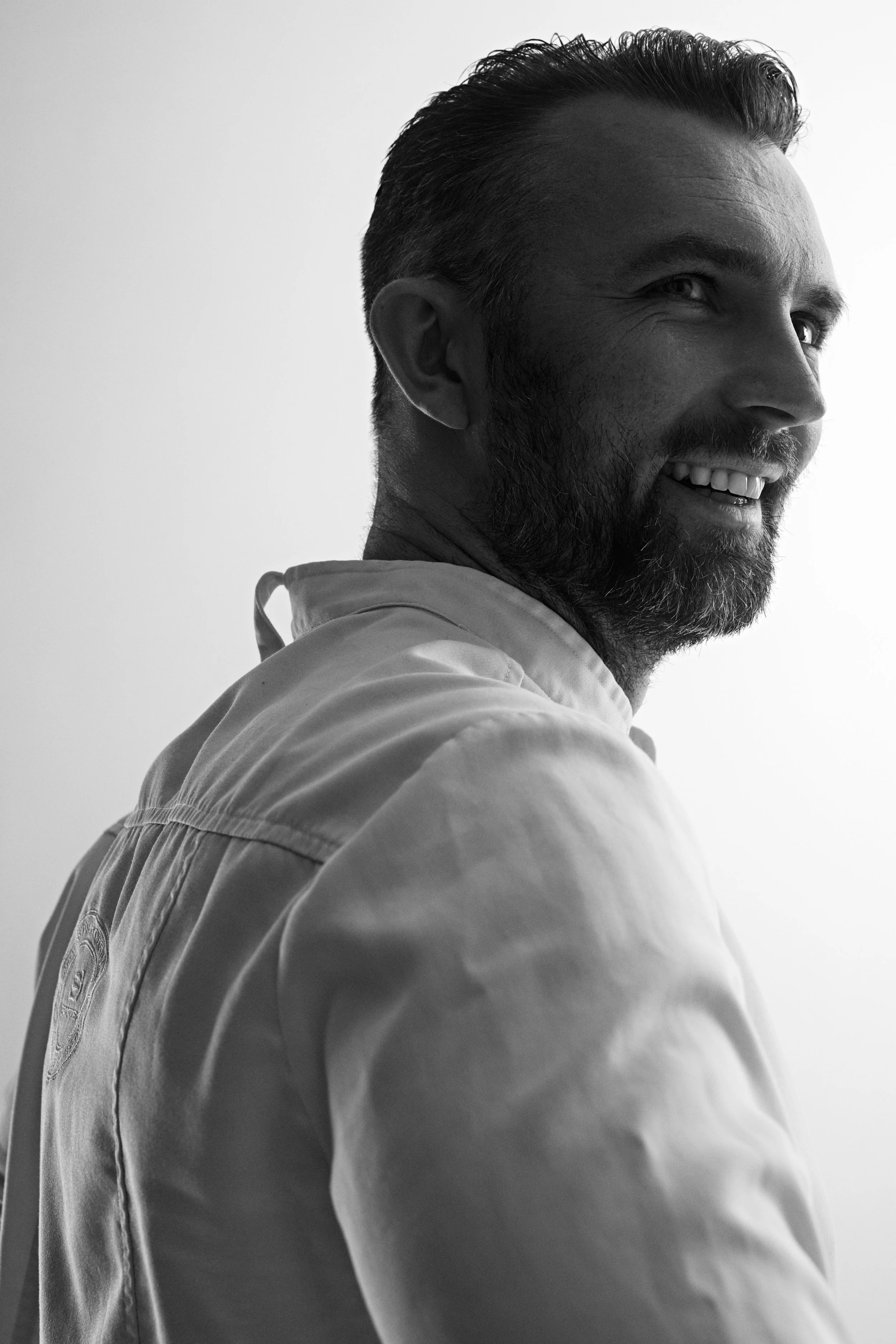 Black and white photo of a man smiling, looking to the side, with short hair and a beard, wearing a collared shirt.