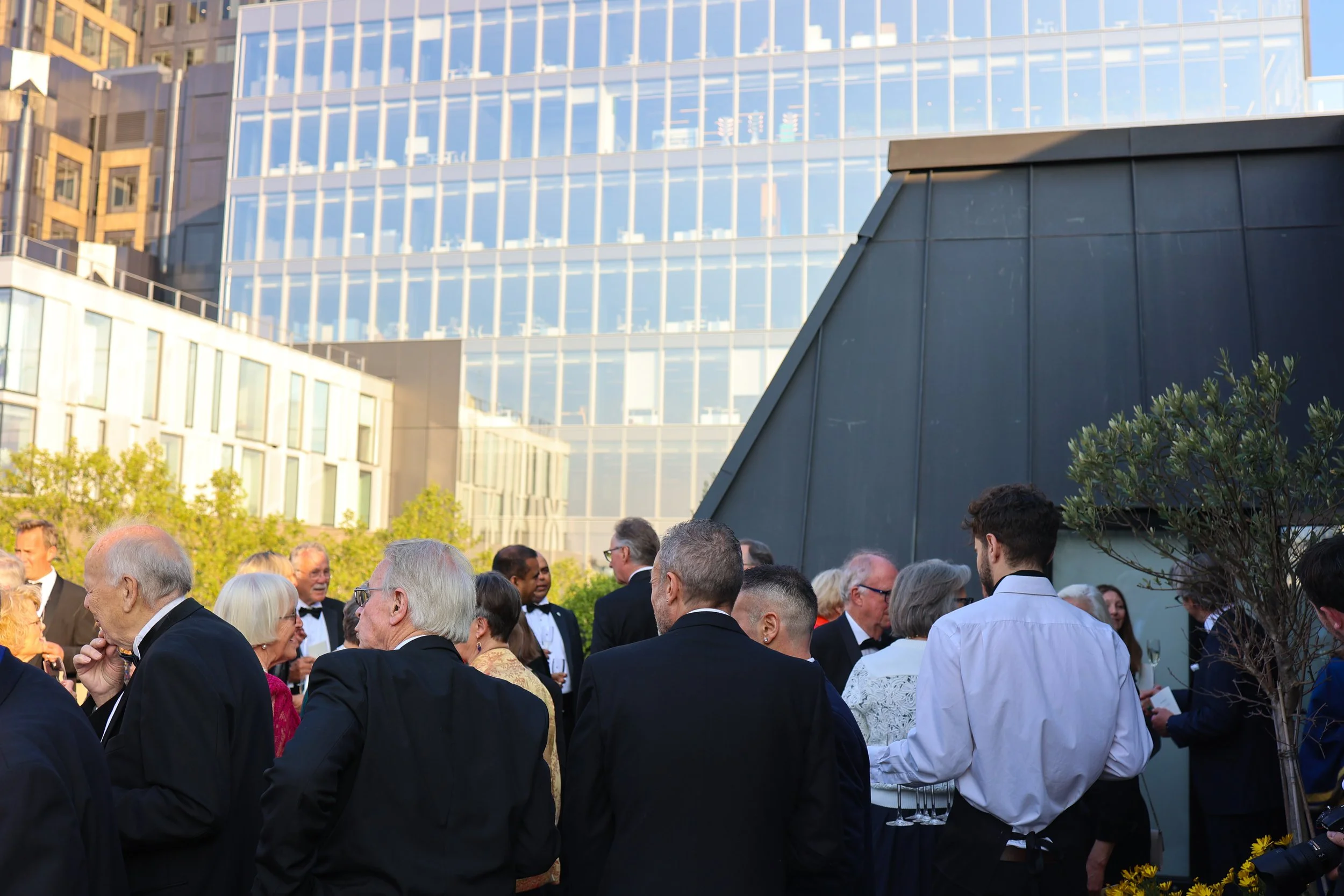 A group of people in formal attire gathered outdoors in front of modern glass buildings during a sunny day.