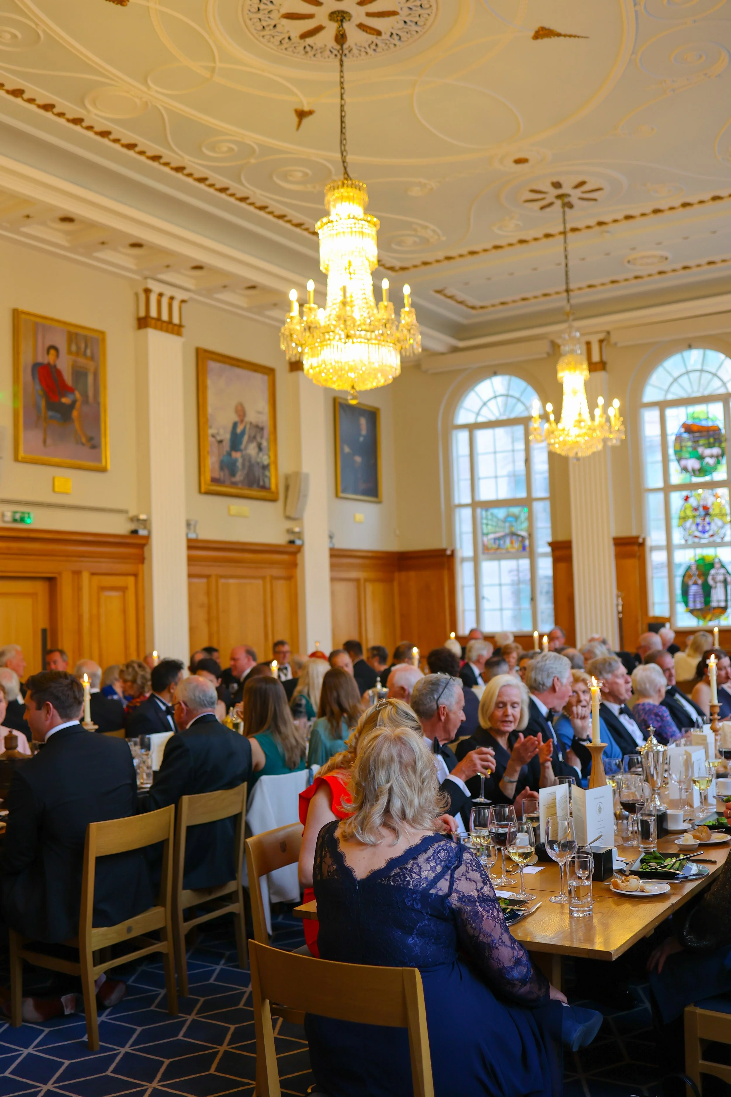 A large banquet hall filled with formally dressed people sitting at long tables. The hall features ornate chandeliers hanging from a decorated ceiling, large arched windows with stained glass, wood-paneled walls, and framed portraits.