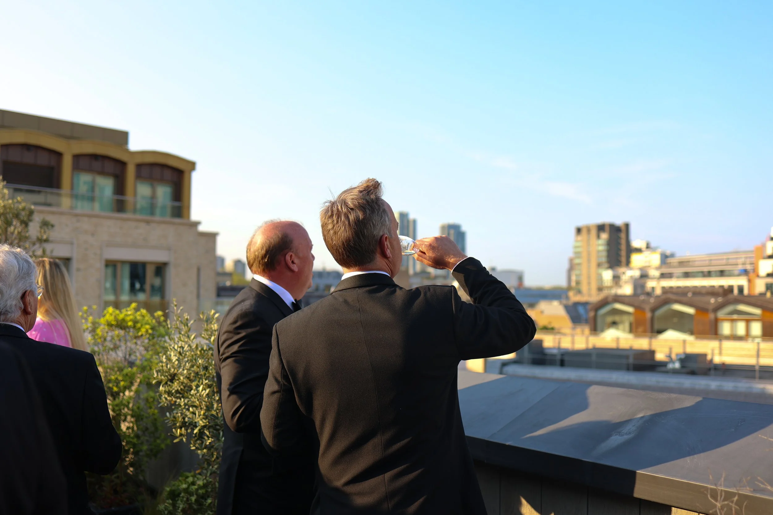 A group of people on a rooftop terrace, with a city skyline in the background, dressed in formal attire, some holding drinks, during sunset.