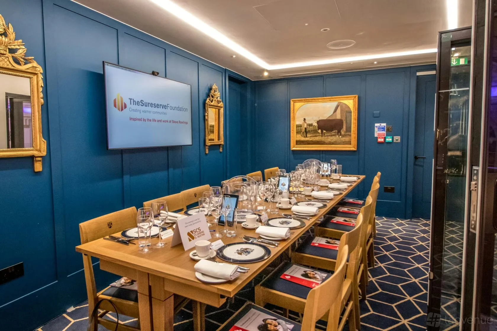 A long dining table set with white plates, wine glasses, water glasses, cups, and napkins in a room with blue walls, gold framed mirrors, and paintings, preparing for a meeting or dinner event.