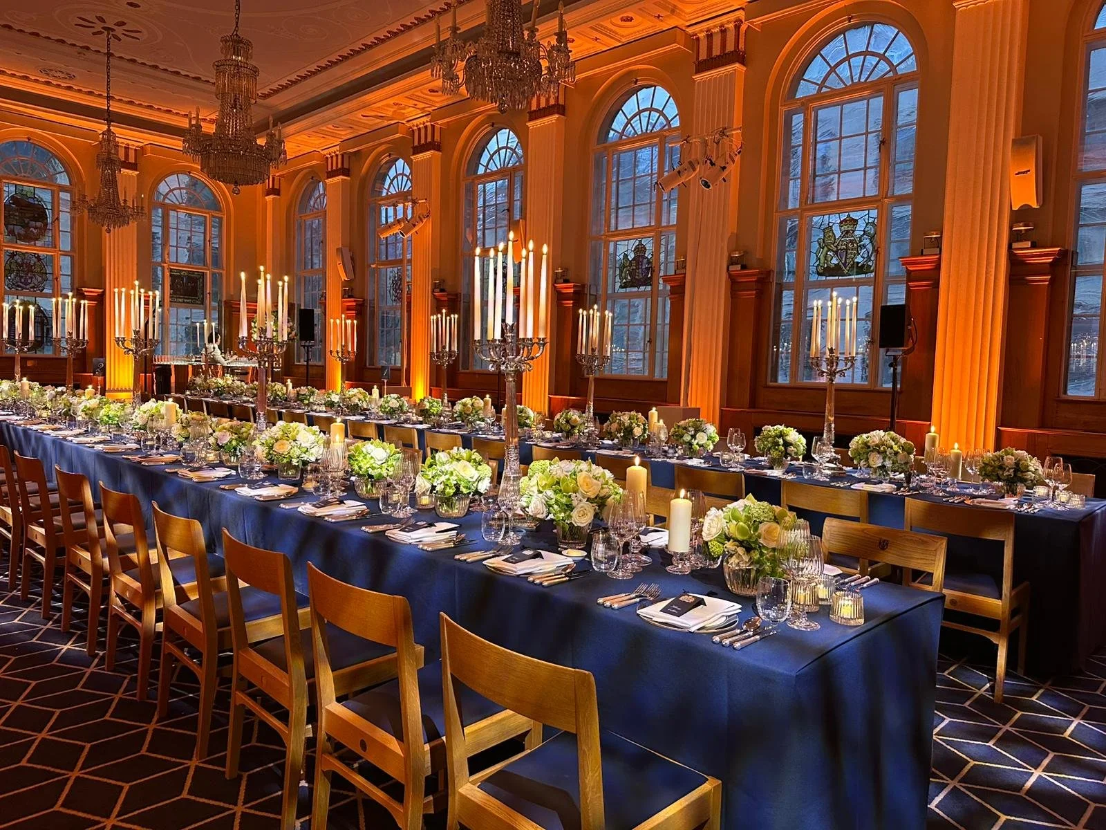 Elegant banquet hall decorated with tall candelabras, floral arrangements, and candles on a long dining table, with high arched windows and chandeliers in the background.