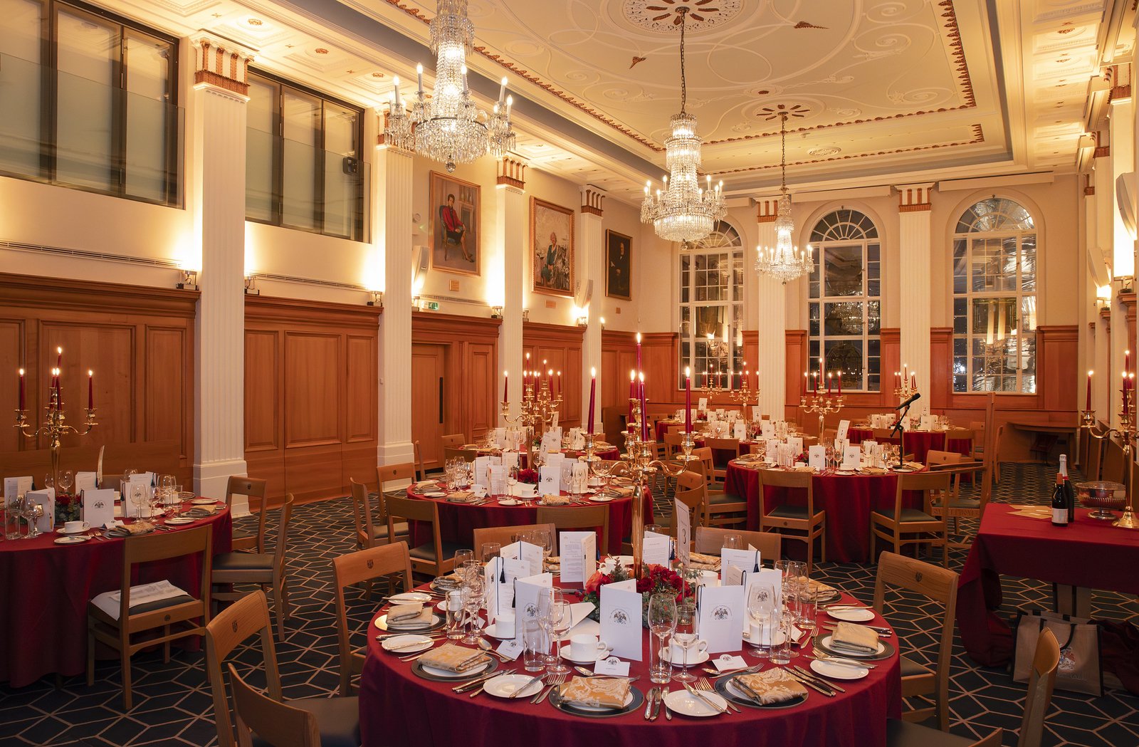 A finely decorated banquet hall with round tables covered in red tablecloths, elegant place settings, and tall candlesticks. The room has large arched windows, wooden-paneled walls, ornate chandeliers, and framed portraits on the walls.
