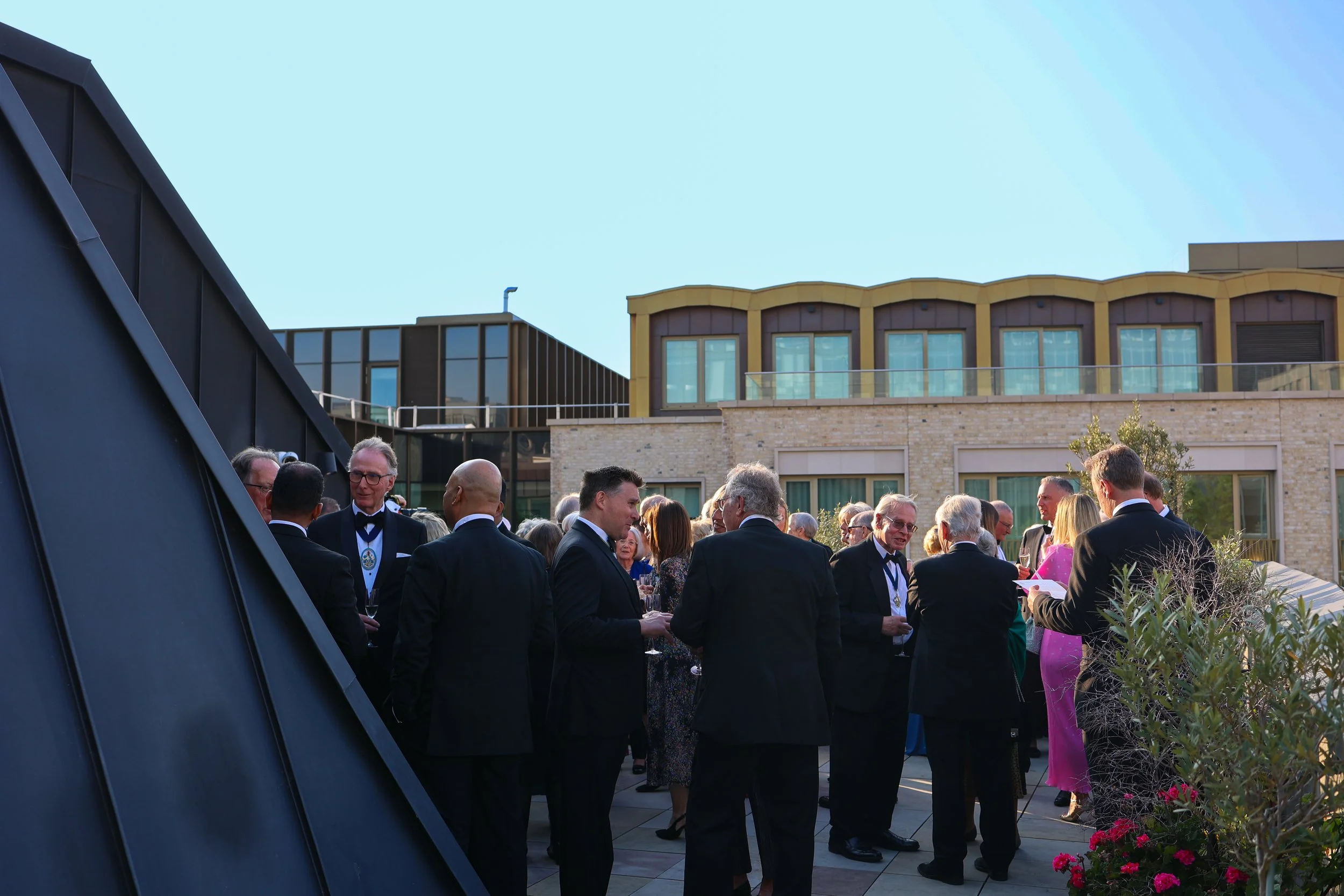 A group of people dressed in formal attire attending an outdoor wedding or celebration on a terrace with modern buildings in the background.