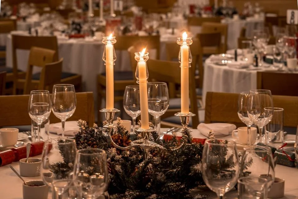 Elegant banquet table decor featuring three lit white candles on silverholders, surrounded by pine branches and berries, set in a dimly lit event hall with multiple tables laid out for a celebration.