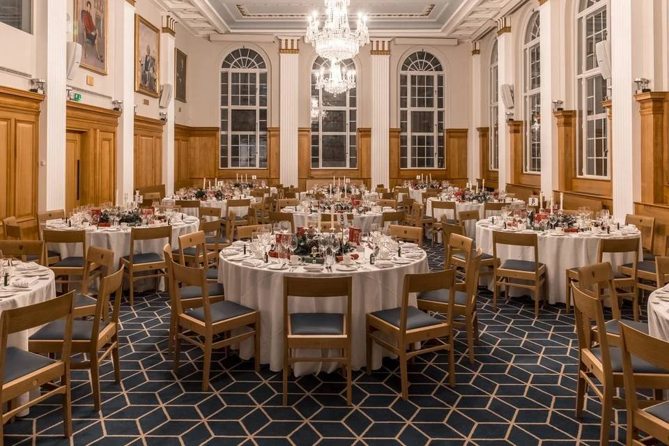 Elegant banquet hall decorated for a formal event with round tables covered in white tablecloths, set with glassware and centerpieces, wooden chairs with dark cushions, large arched windows, high ceilings with chandeliers, and framed paintings on the